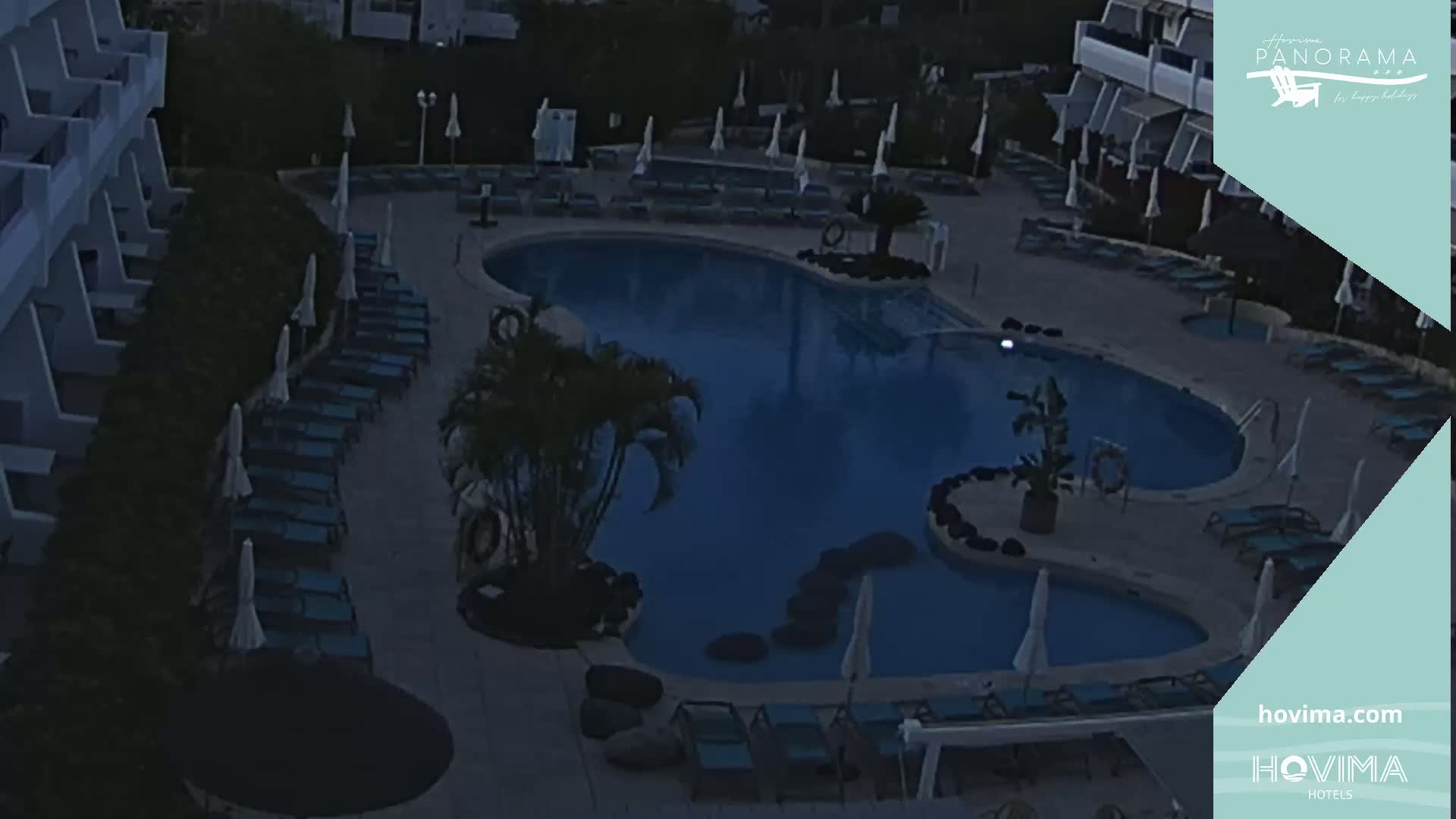 An outdoor swimming pool at night, surrounded by lounge chairs and umbrellas, is next to a building with balconies.
