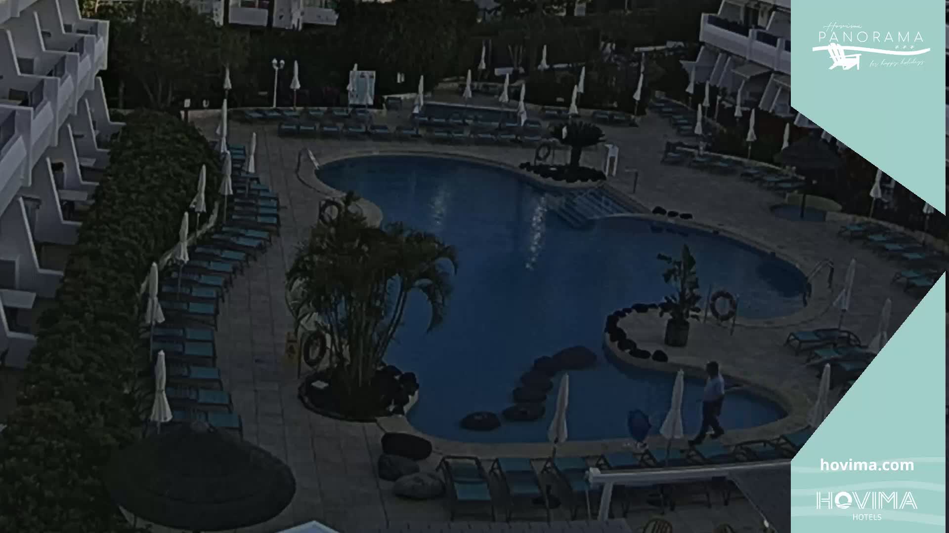 An outdoor swimming pool area at dusk, with rows of lounge chairs and umbrellas surrounding a curving pool, next to a building.