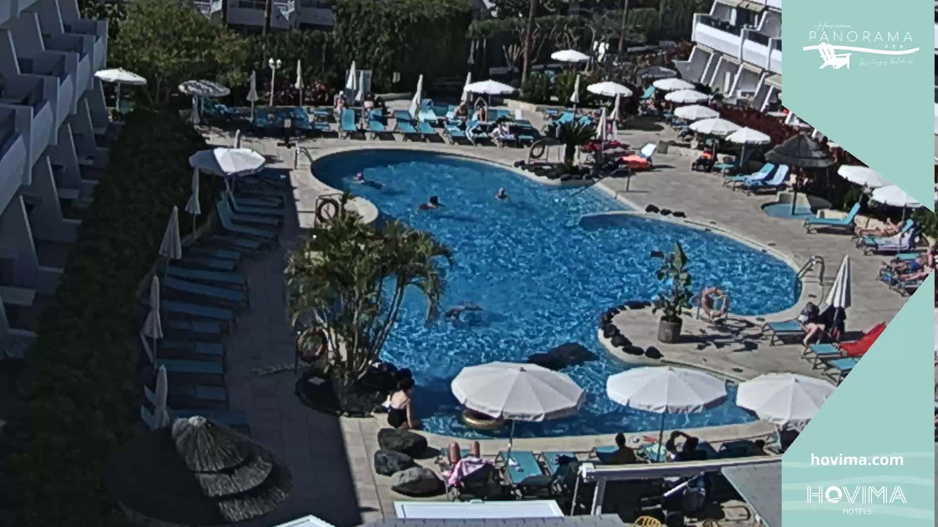 An outdoor pool surrounded by lounge chairs, umbrellas, and a building under sunny conditions.