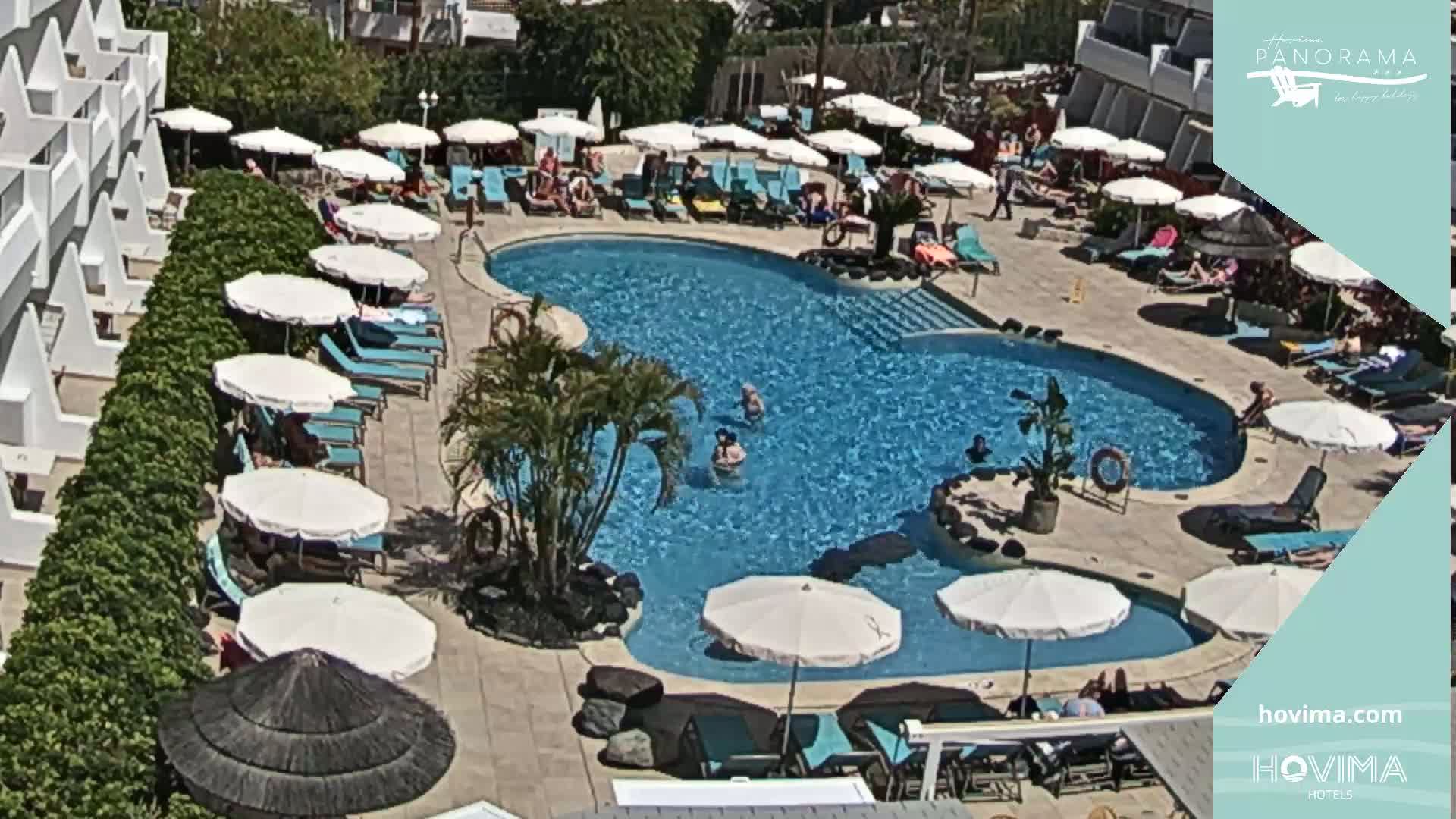 A sunny outdoor scene shows a kidney-shaped swimming pool surrounded by lounge chairs, umbrellas, and a building.