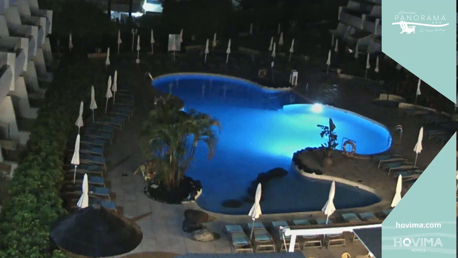 An illuminated, kidney-shaped swimming pool at night surrounded by lounge chairs and umbrellas, next to a building.