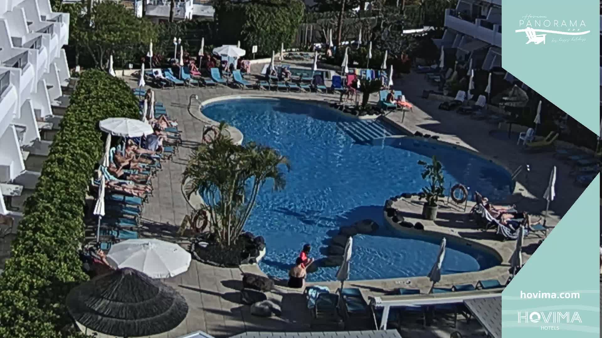 An aerial view shows a sunny resort pool area with numerous people lounging on chairs and in the water surrounding a kidney-shaped pool.