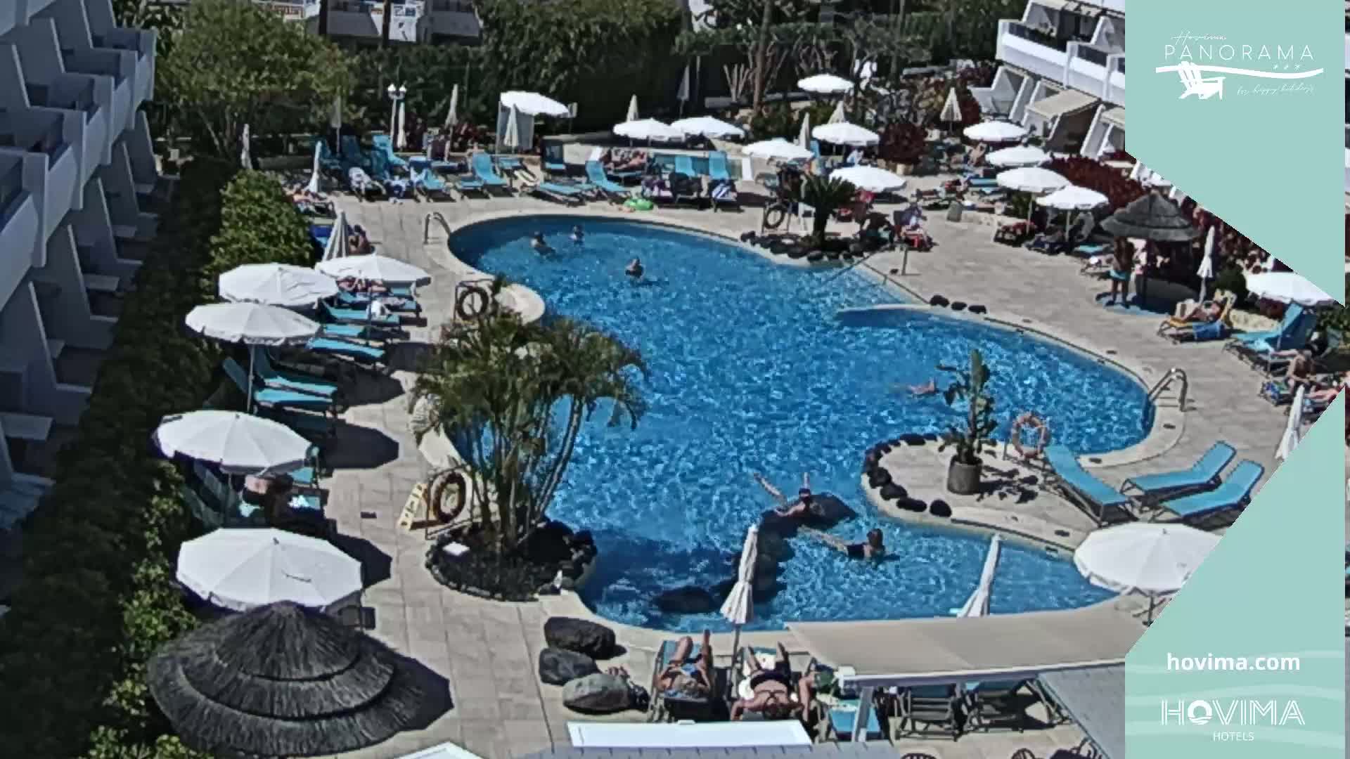 An aerial view shows a resort pool area with numerous blue lounge chairs and white umbrellas surrounding a large, irregularly shaped swimming pool on a sunny day.