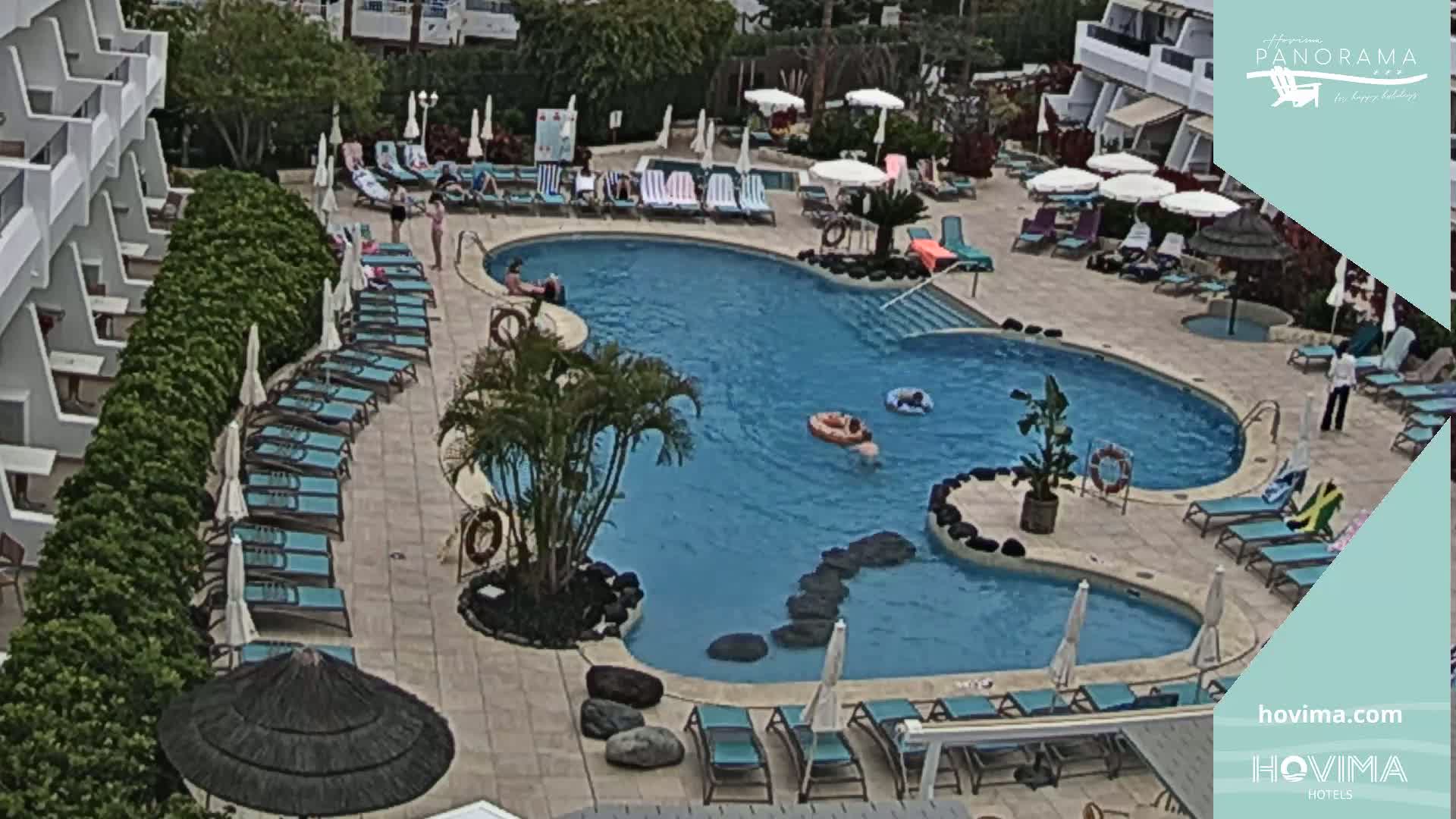 An outdoor pool surrounded by lounge chairs and umbrellas at a resort, with several people in and around the pool on a partly cloudy day.