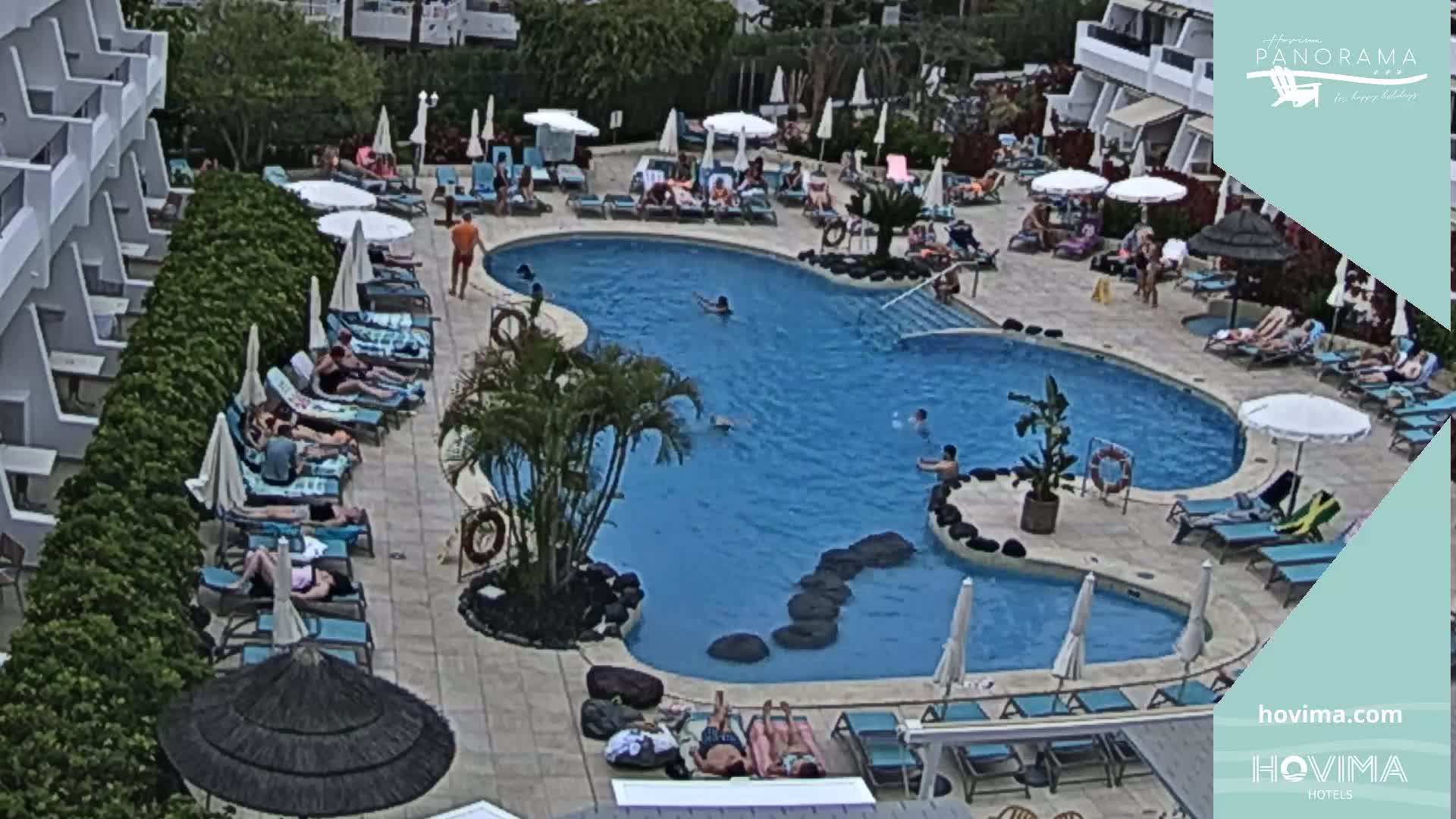 An outdoor resort pool area with many people lounging on chairs and in the water on a partly cloudy day.