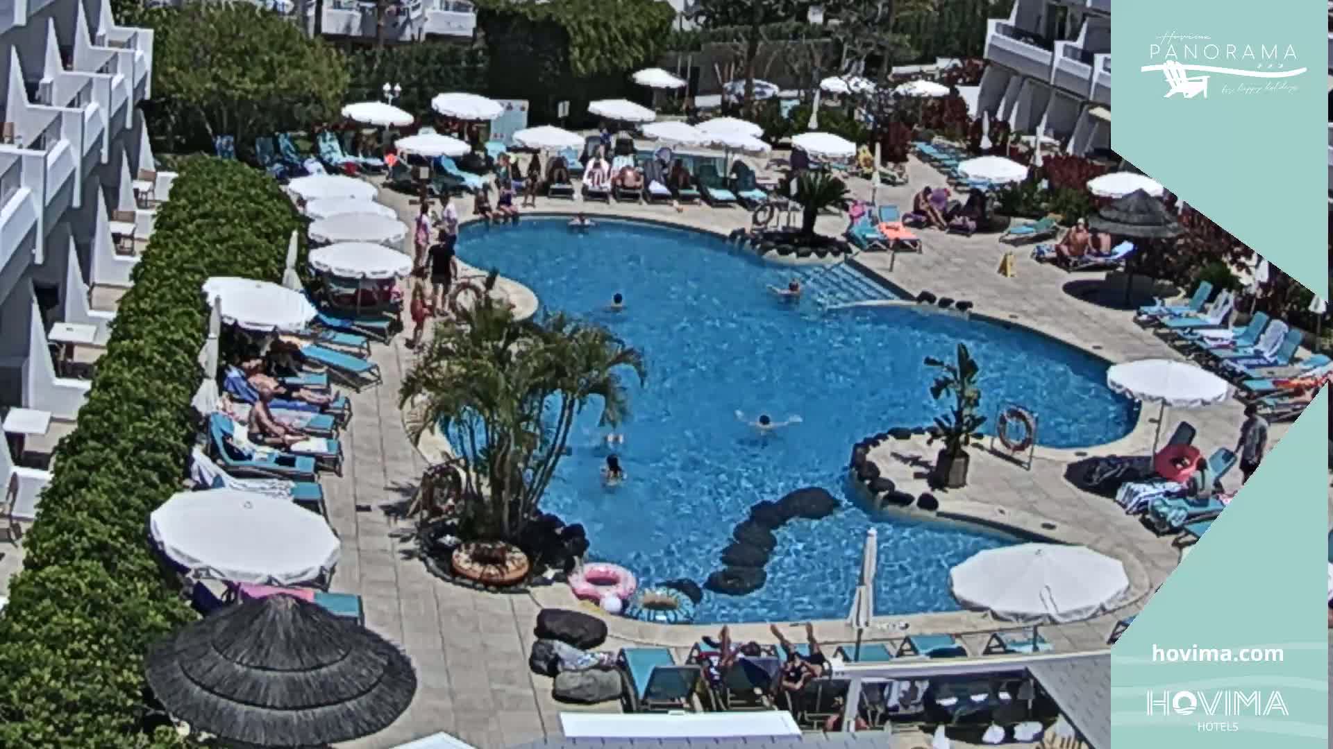 An outdoor pool surrounded by lounge chairs, umbrellas, and a building, under sunny skies.