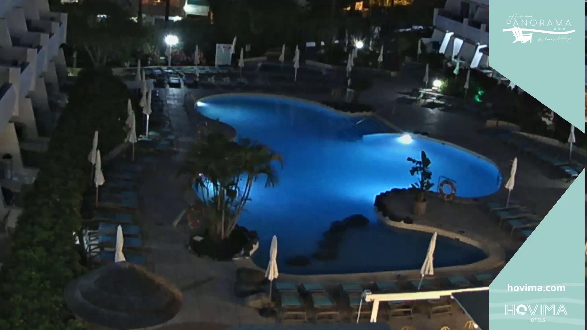 An illuminated, kidney-shaped swimming pool surrounded by lounge chairs and umbrellas at night.