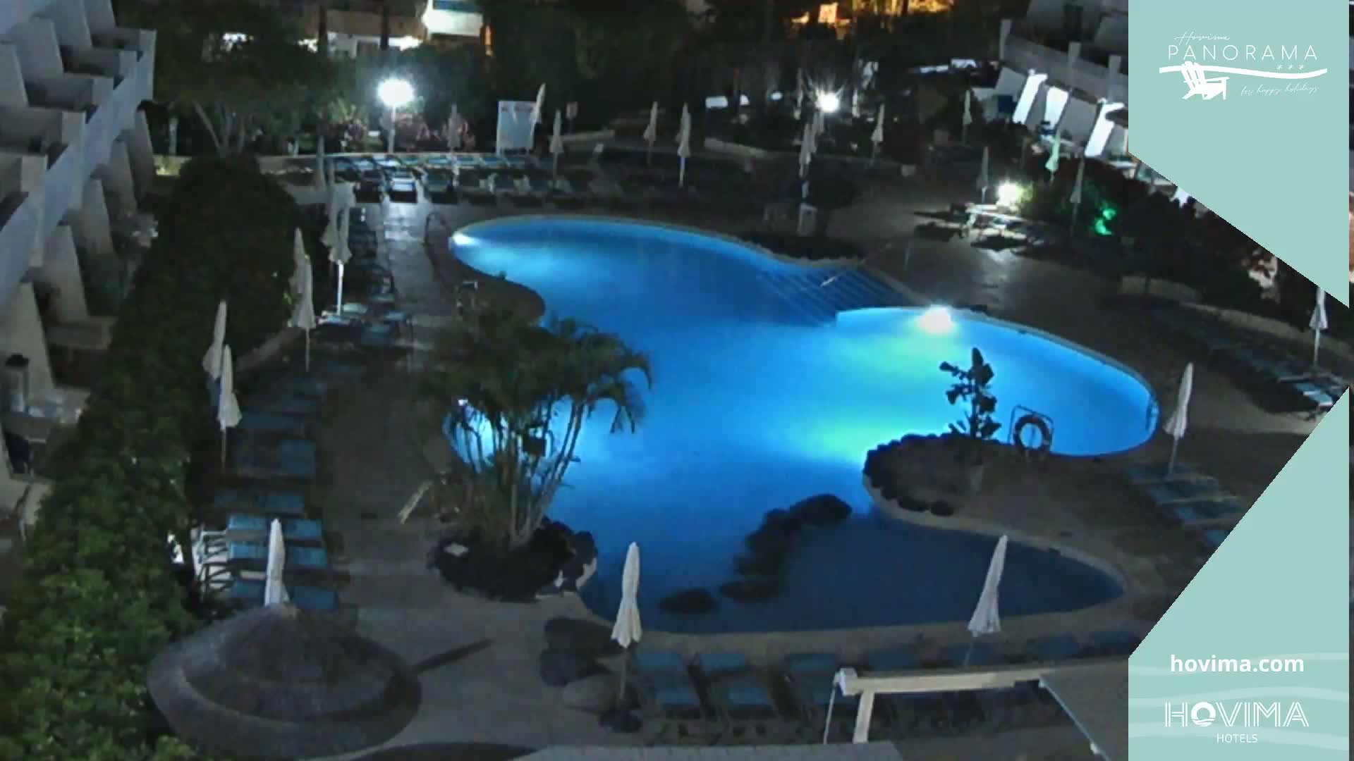 A brightly lit, kidney-shaped swimming pool at night, surrounded by lounge chairs and palm trees near a building.
