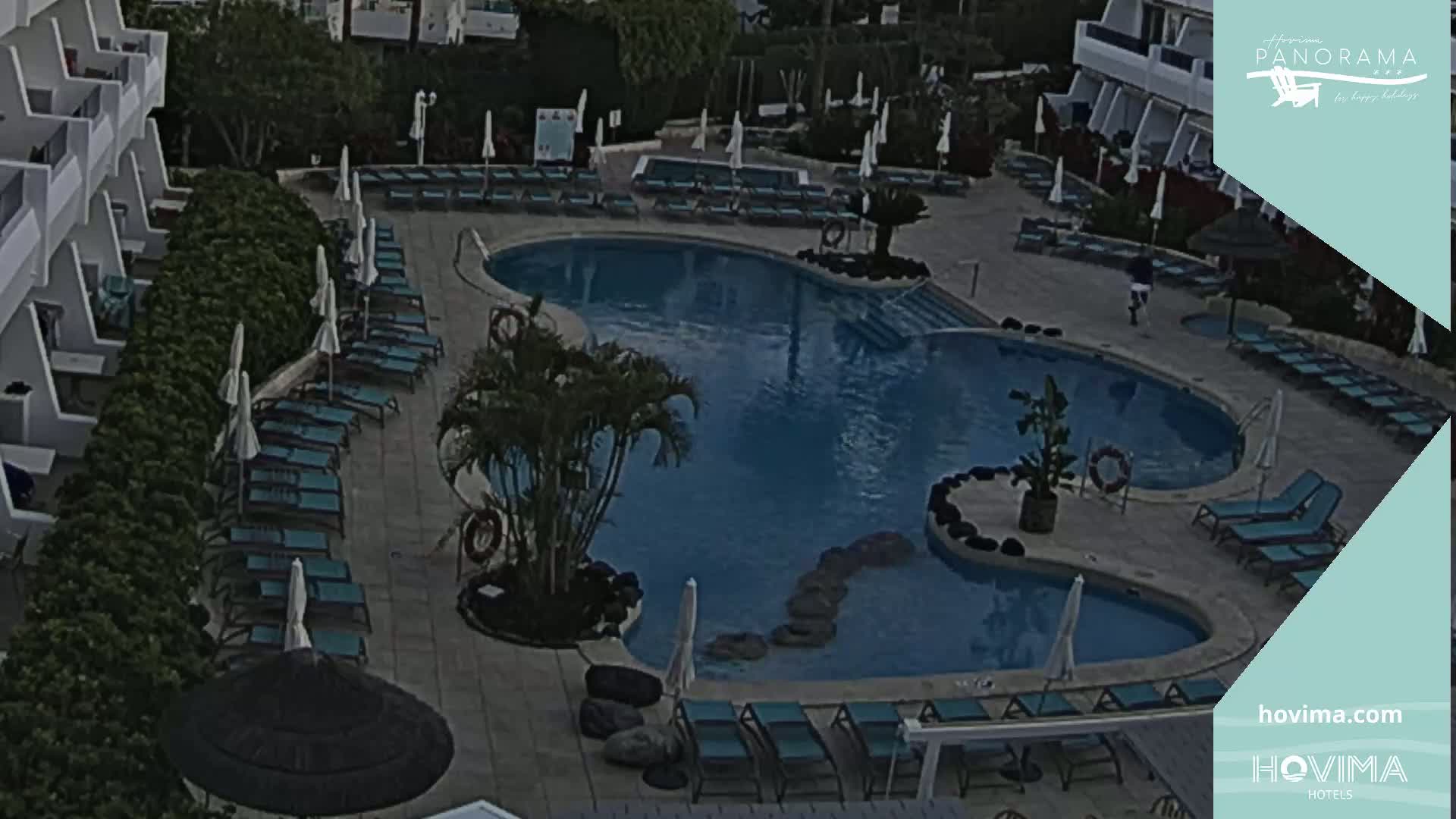 An overhead view shows a swimming pool surrounded by lounge chairs and umbrellas at a resort, with a person walking near the pool.