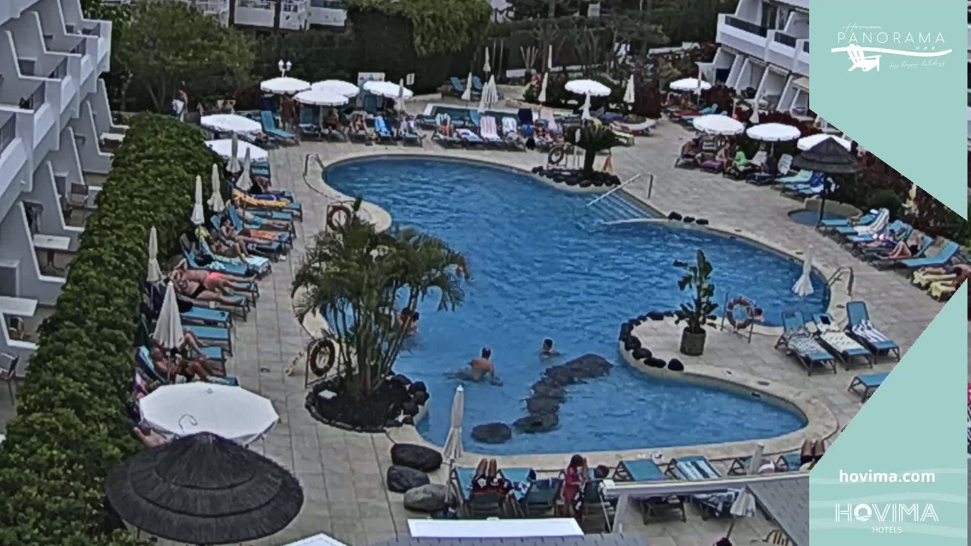 An outdoor pool surrounded by lounge chairs and umbrellas at a resort, with people swimming and sunbathing under a partly cloudy sky.