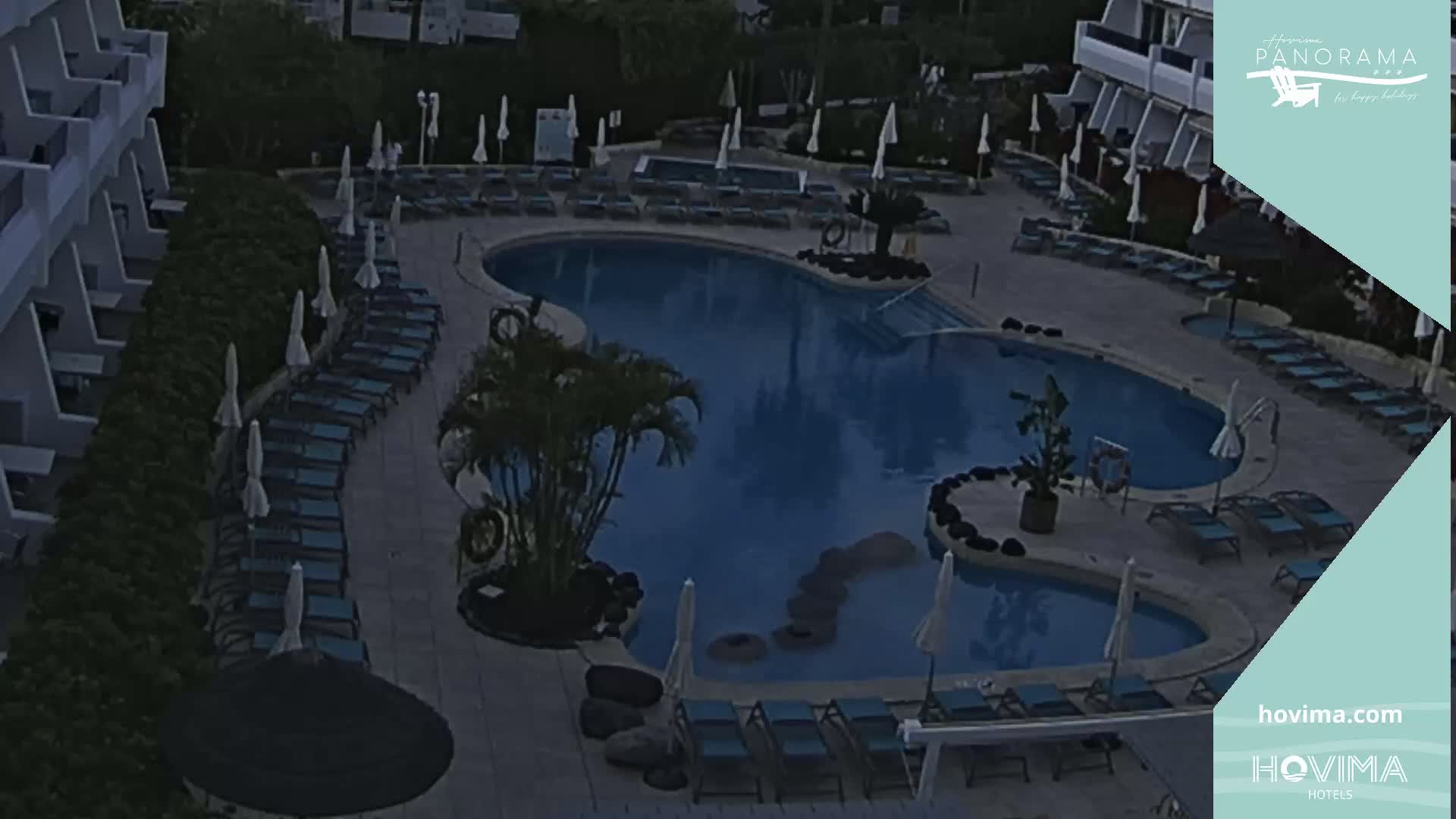 An outdoor swimming pool at dusk, surrounded by lounge chairs and umbrellas, is nestled between a building and lush vegetation.