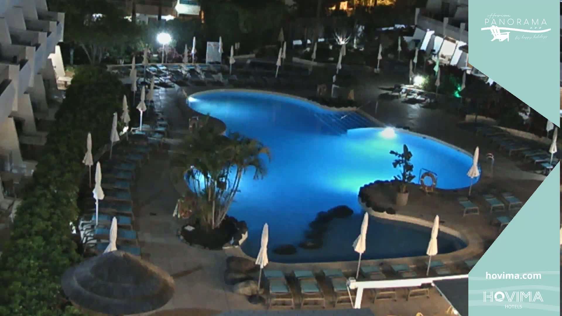 A nighttime view of a resort pool area with lounge chairs and umbrellas surrounding a brightly lit blue pool.