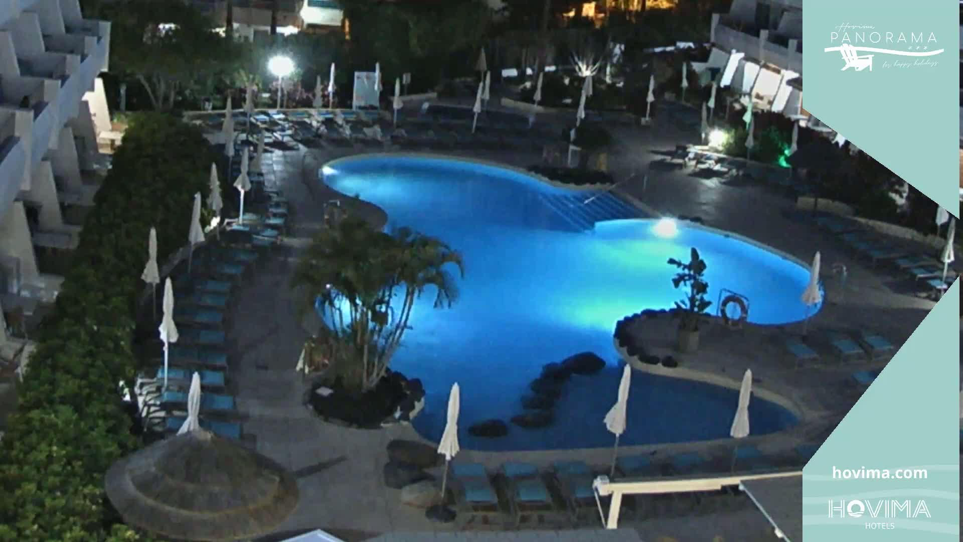 A brightly lit, irregularly shaped swimming pool at night is surrounded by lounge chairs and umbrellas near a building.
