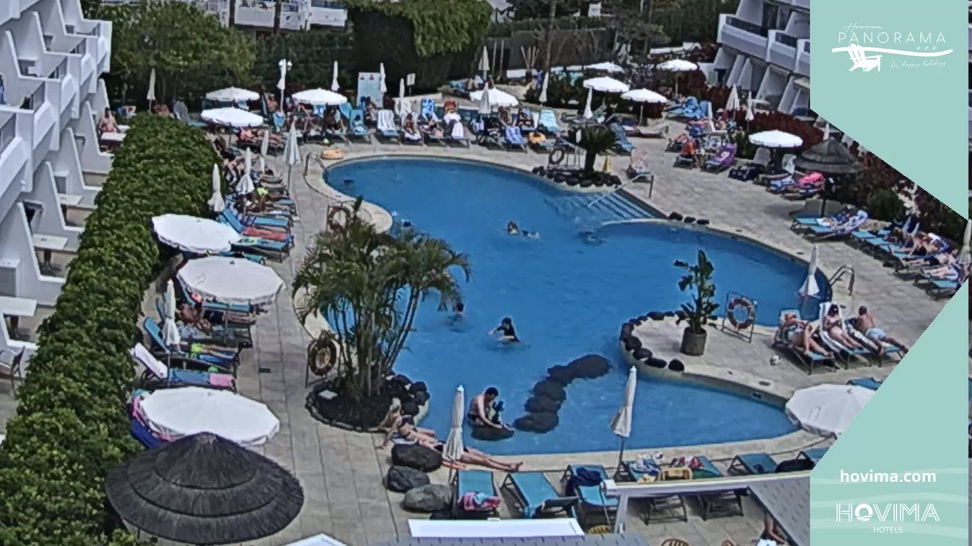 A resort pool area with numerous people swimming, sunbathing, and lounging under umbrellas on a sunny day.