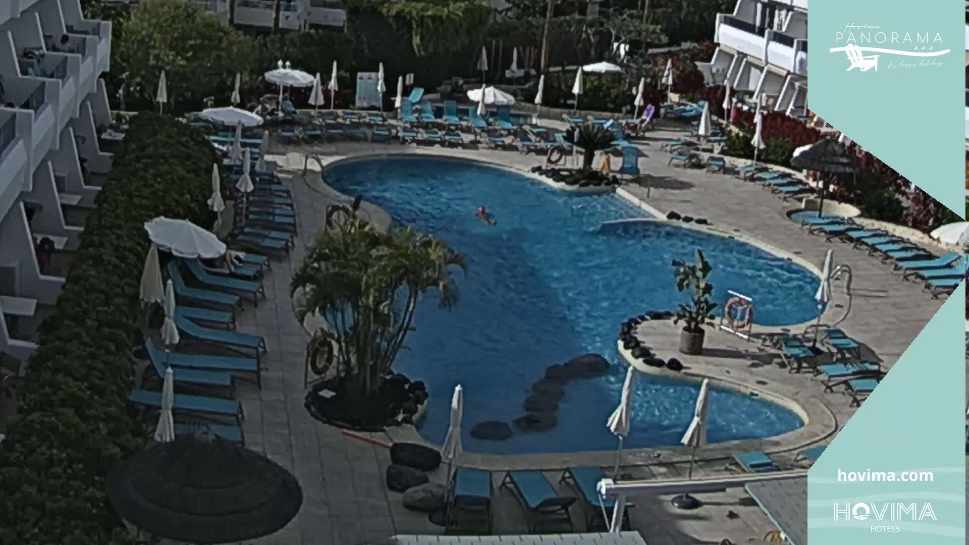 An outdoor pool with surrounding lounge chairs and umbrellas, next to a building, under a sunny sky.