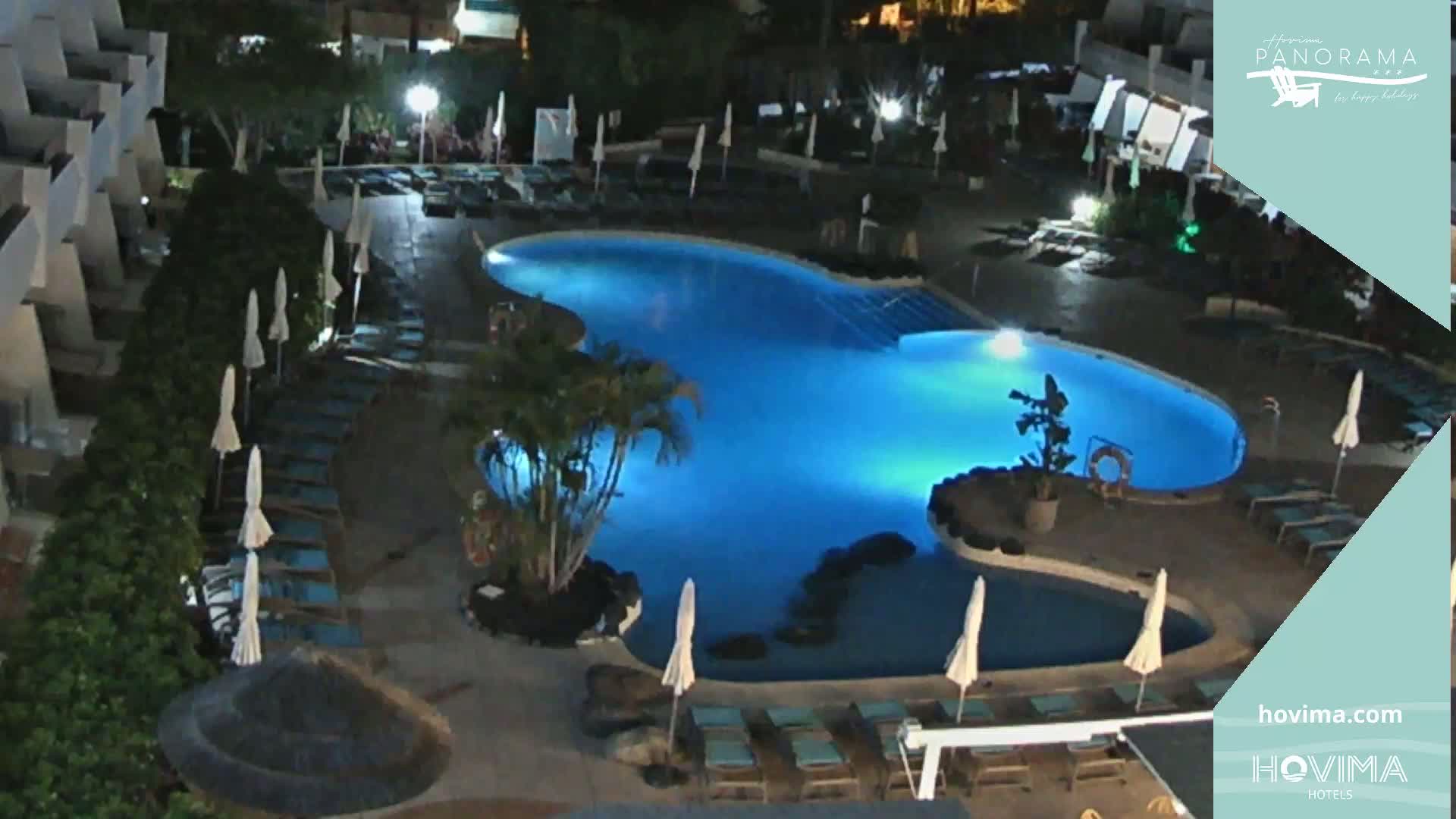An illuminated, kidney-shaped swimming pool surrounded by lounge chairs and umbrellas at night.