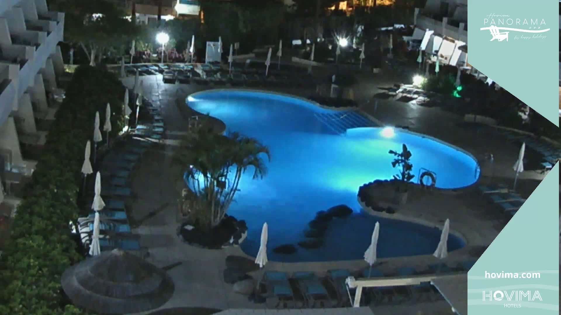 A brightly lit, kidney-shaped swimming pool at night is surrounded by lounge chairs and umbrellas near a building.