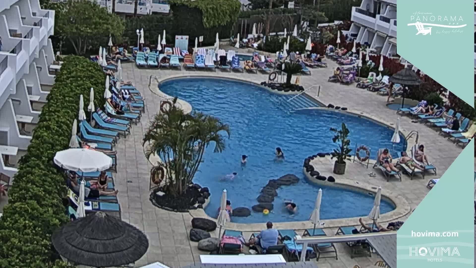 An outdoor swimming pool surrounded by lounge chairs and umbrellas, with people swimming and sunbathing on a partly sunny day.