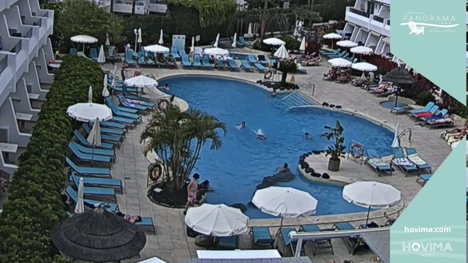 An outdoor pool surrounded by lounge chairs and umbrellas, with people swimming and sunbathing in sunny weather.