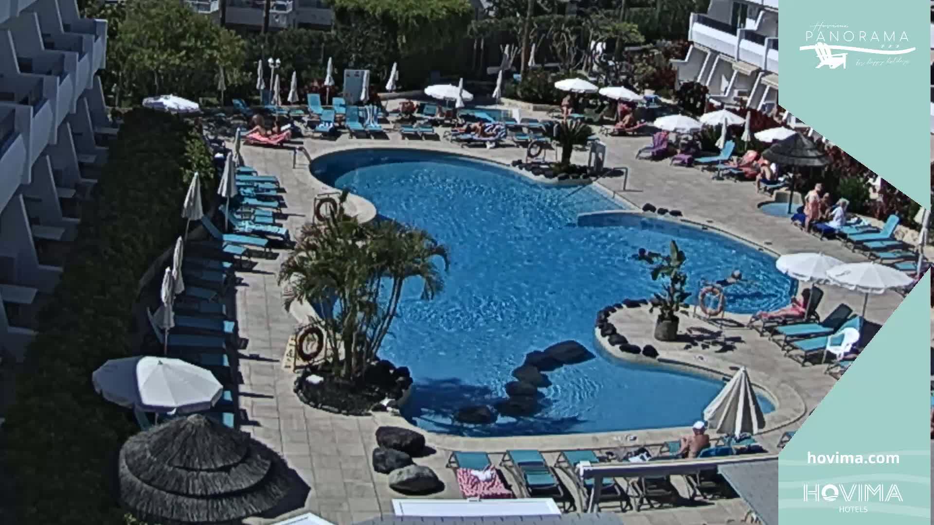 A sunny outdoor scene shows a large kidney-shaped swimming pool surrounded by lounge chairs, umbrellas, and people relaxing.