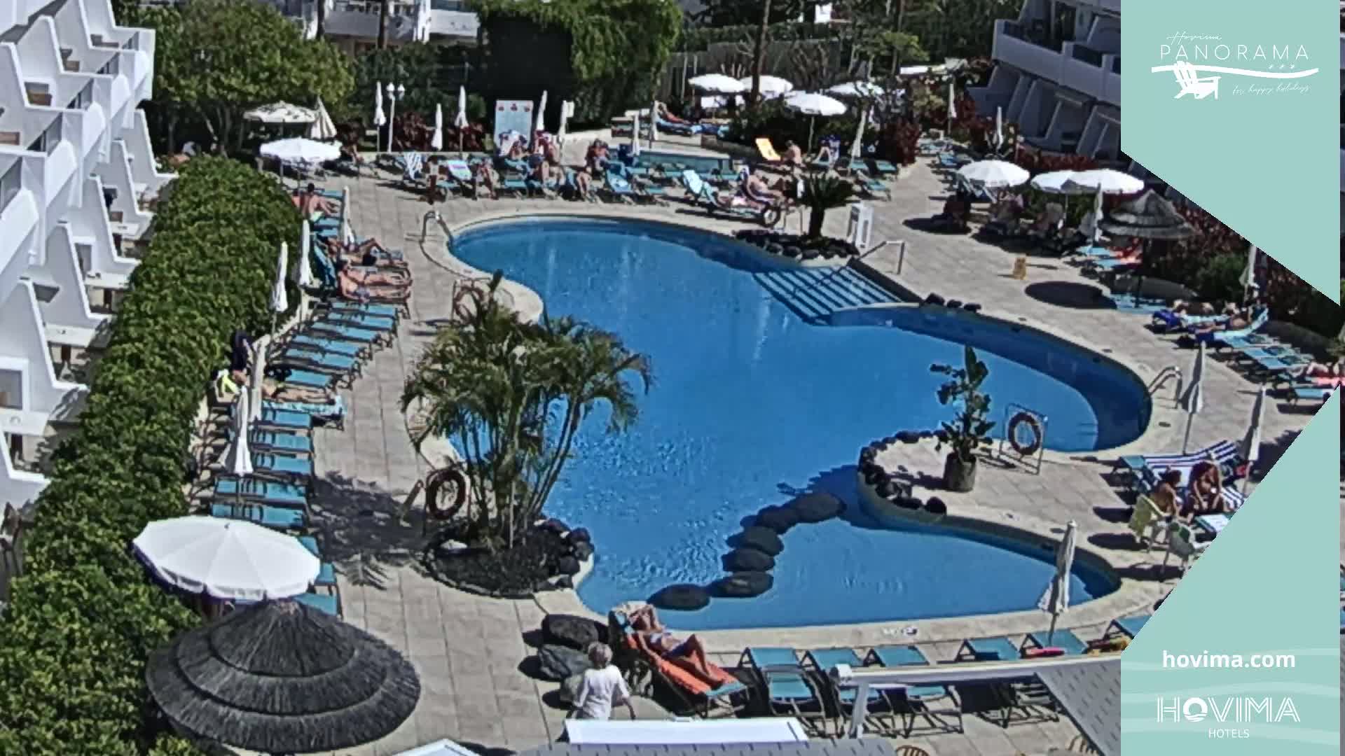 A sunny outdoor scene shows a kidney-shaped swimming pool surrounded by lounge chairs and umbrellas, adjacent to a white hotel building.