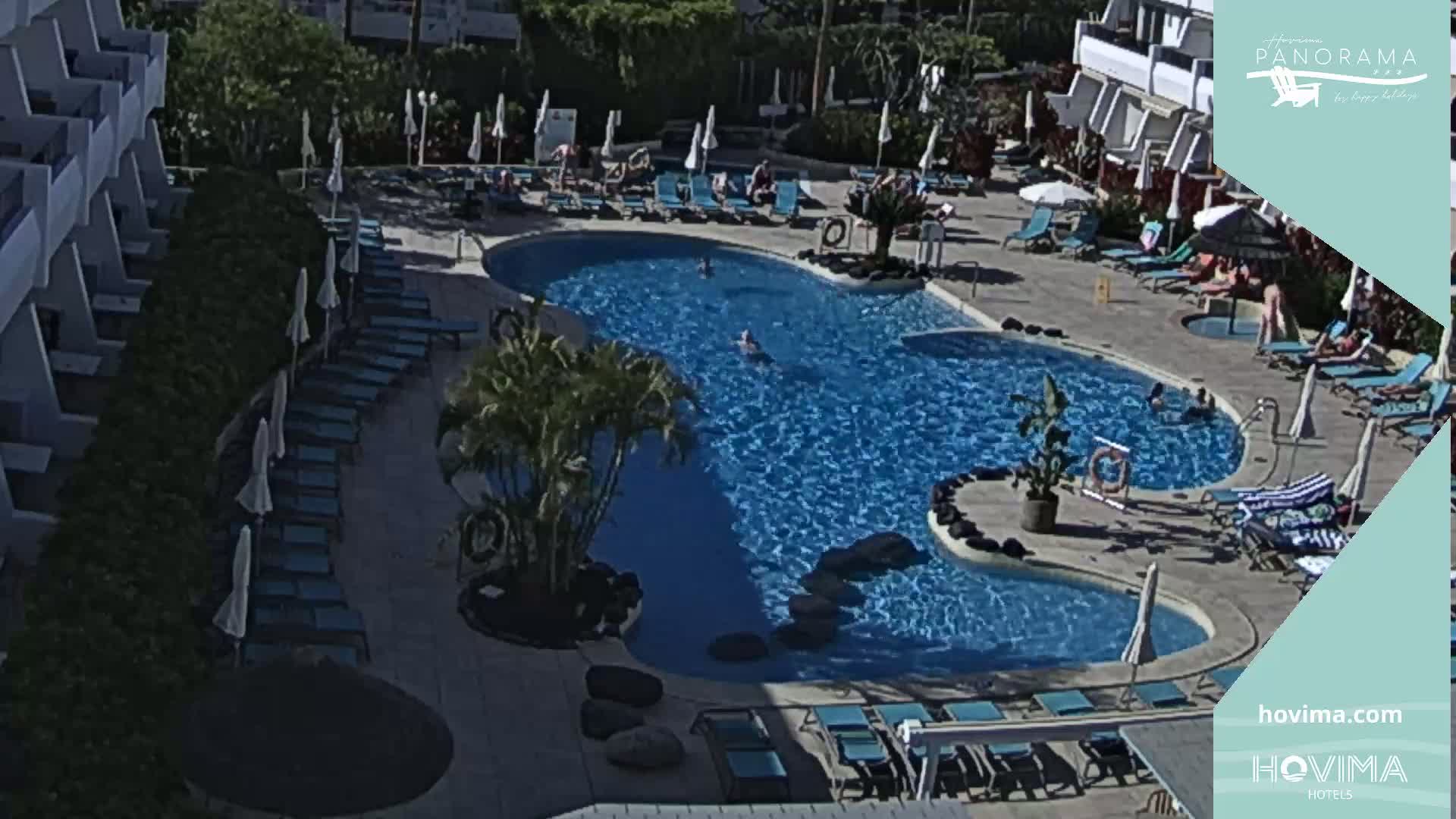 An outdoor pool with lounge chairs and umbrellas surrounds a winding blue pool, partially shaded by buildings and vegetation.