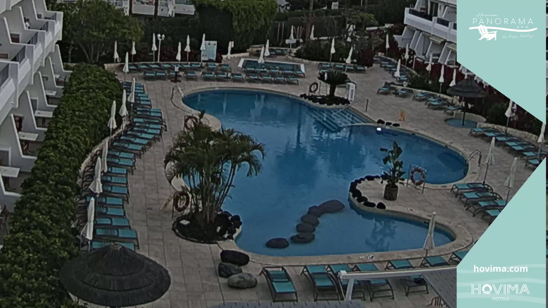 An outdoor pool area with turquoise water, numerous lounge chairs and umbrellas under an overcast sky.