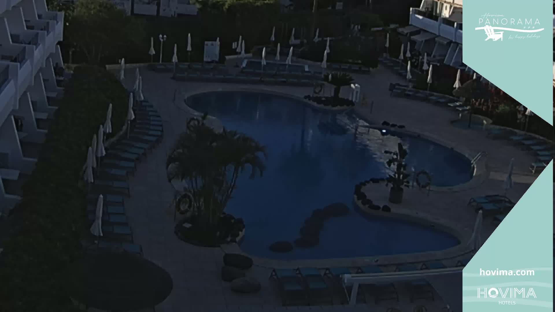 An aerial view shows a resort pool at night, surrounded by lounge chairs and palm trees under a dark sky.