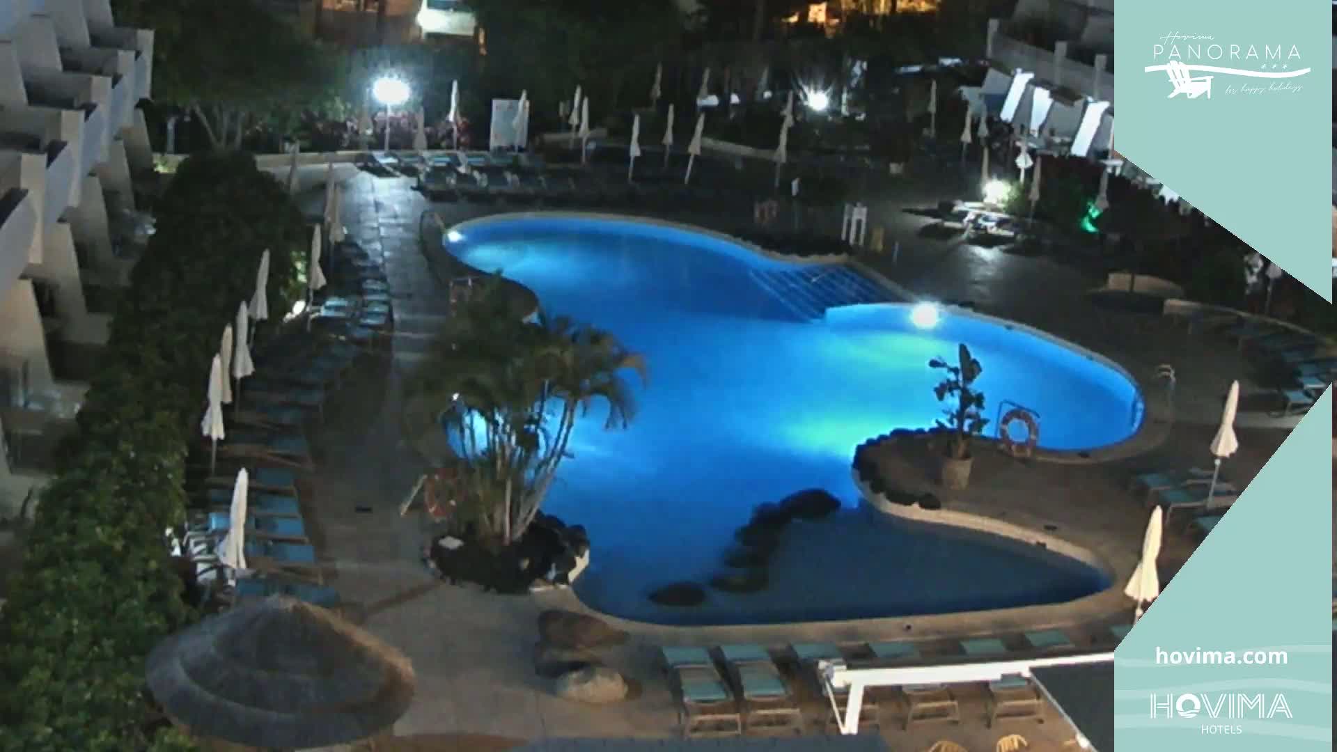 A brightly lit swimming pool at night, surrounded by lounge chairs and palm trees, at a resort.