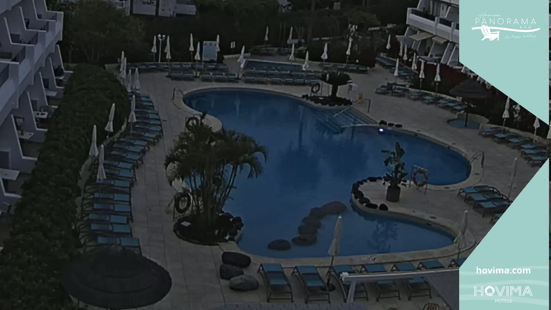 An outdoor swimming pool with surrounding lounge chairs and umbrellas at dusk or night.