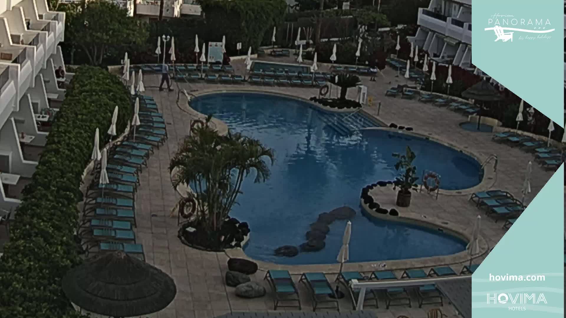 An aerial view shows a kidney-shaped swimming pool surrounded by lounge chairs and umbrellas, next to a building with balconies.