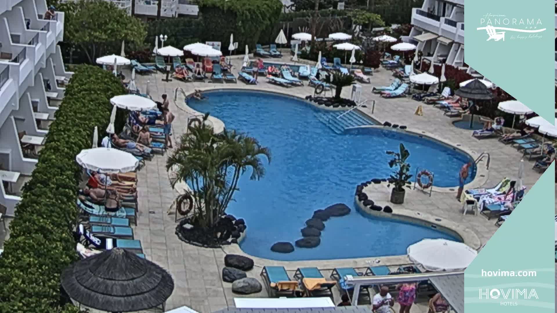 An outdoor pool surrounded by lounge chairs, umbrellas, and a building, with people relaxing in the sunny weather.