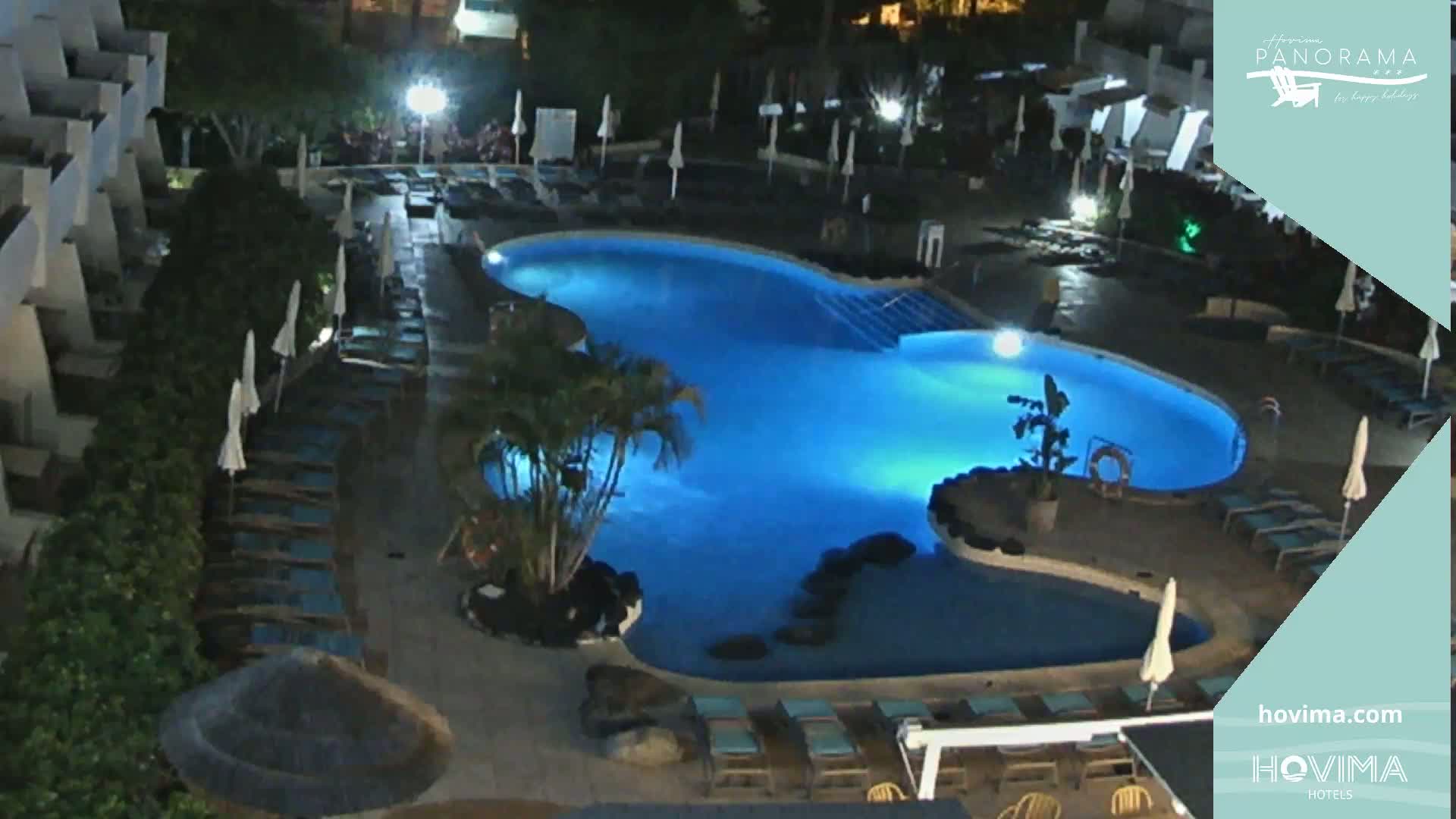 A brightly lit, kidney-shaped swimming pool at night is surrounded by lounge chairs and umbrellas near a building.