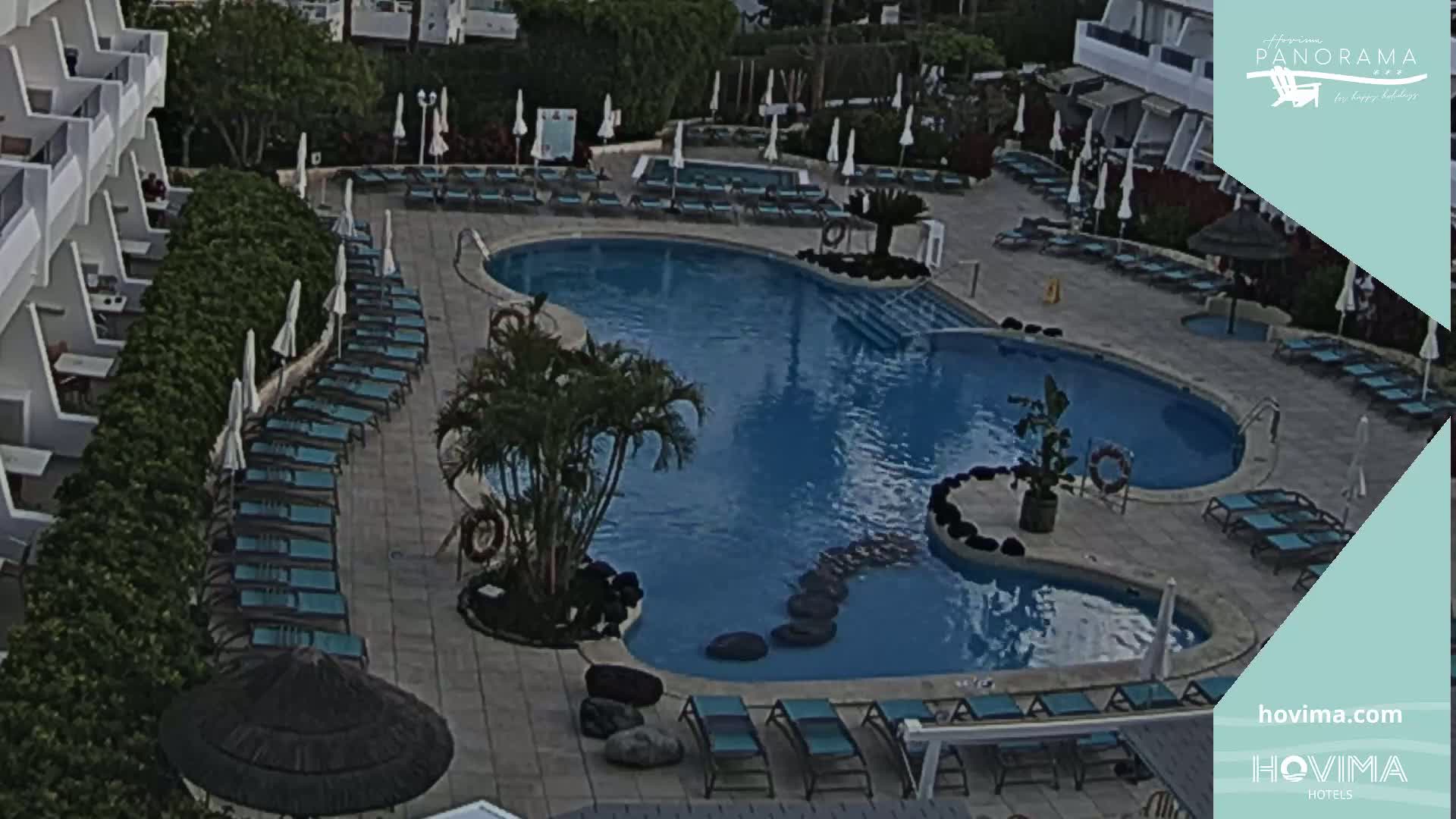 An aerial view shows a resort pool with surrounding lounge chairs and umbrellas under a partly cloudy sky.