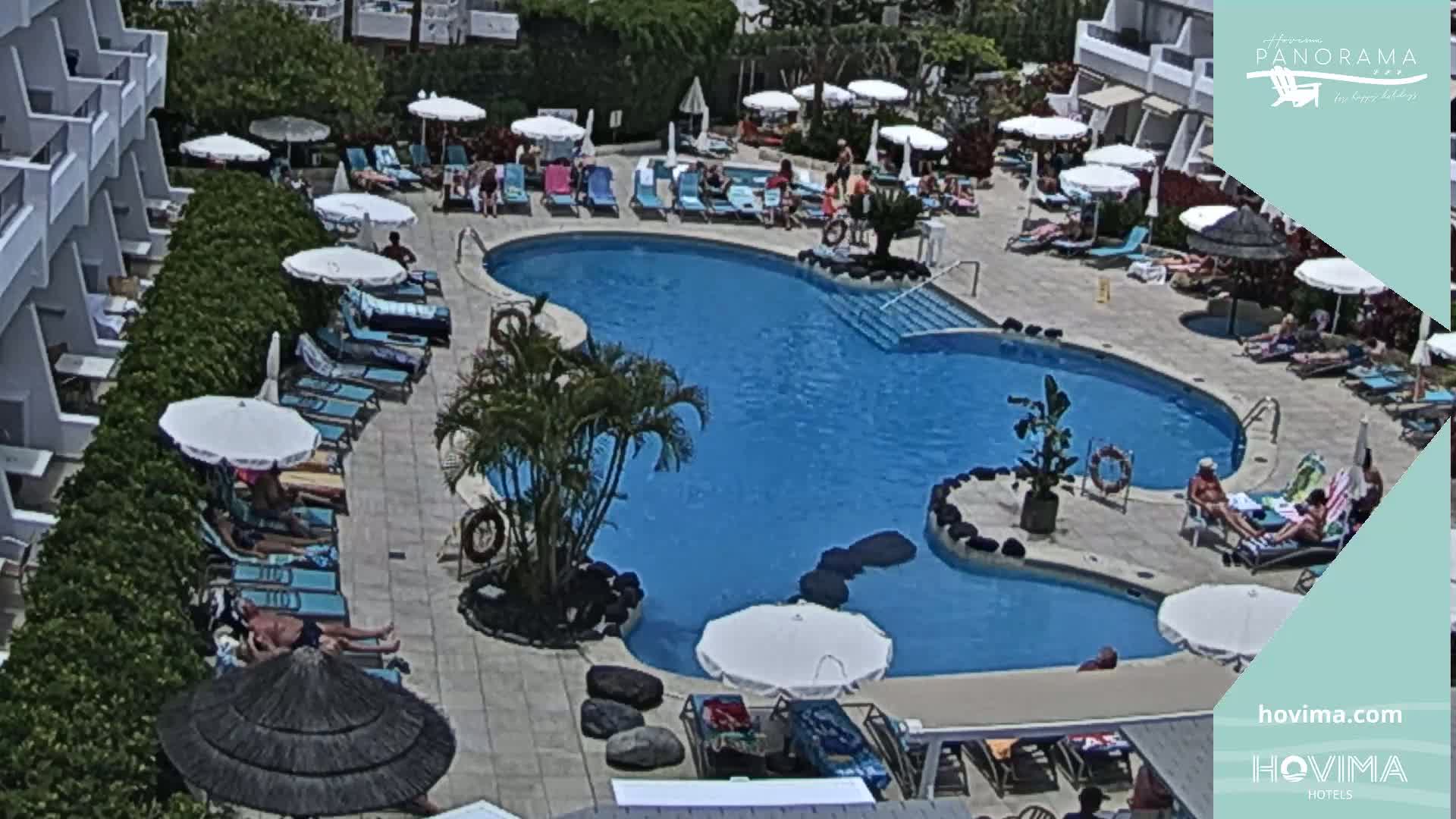 An outdoor swimming pool surrounded by lounge chairs and umbrellas, with people relaxing in the sun on a bright, sunny day.