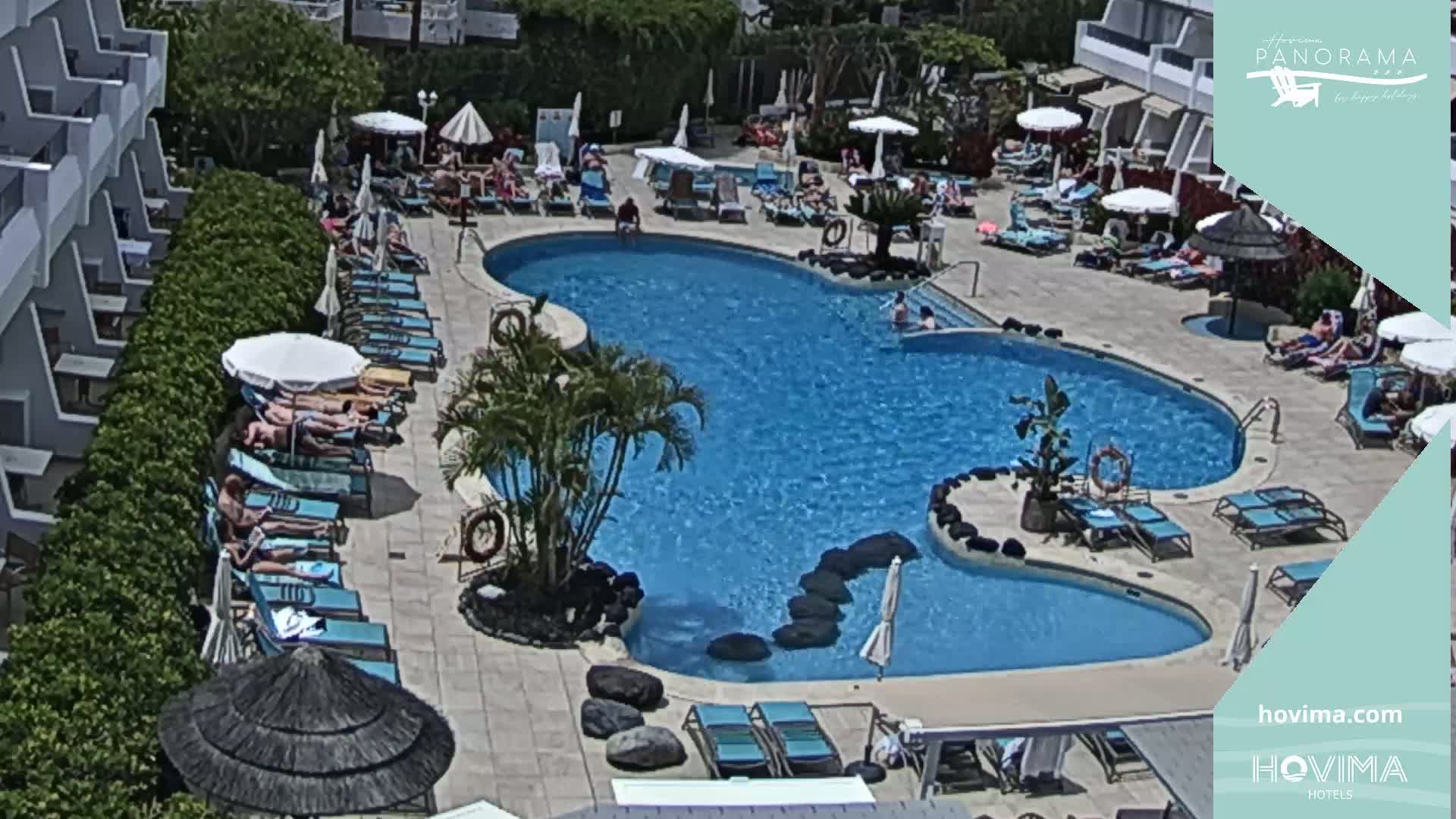 A resort pool area with many people sunbathing on lounge chairs surrounding a kidney-shaped pool on a sunny day.