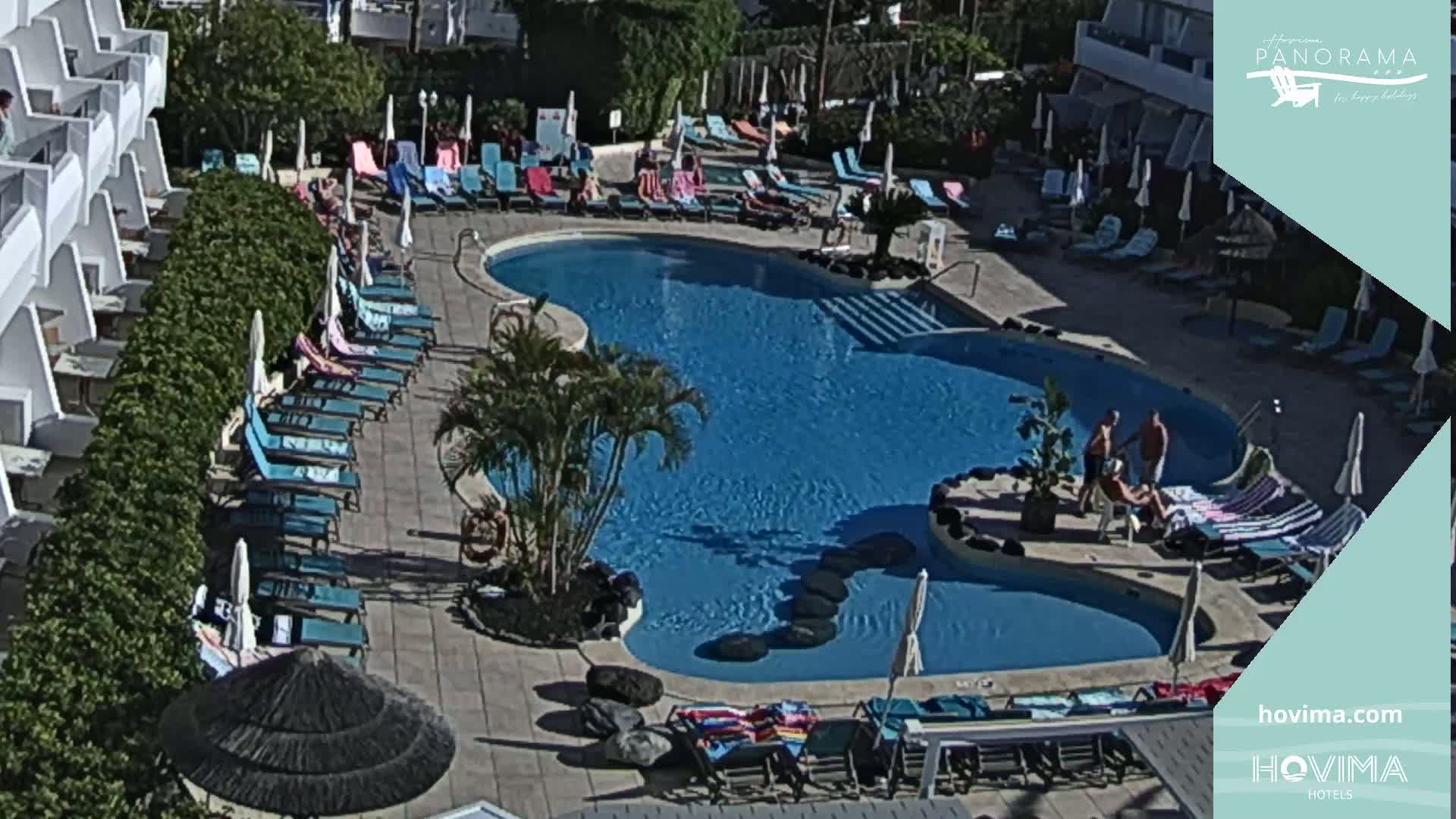 A sunny outdoor scene shows a kidney-shaped swimming pool surrounded by lounge chairs and umbrellas, adjacent to a multi-story building.