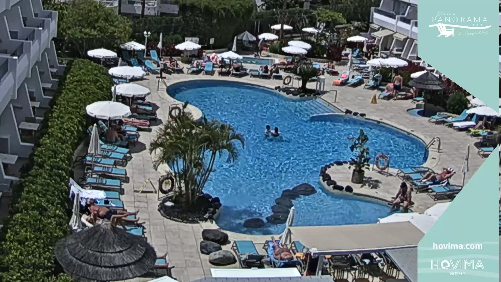 An outdoor pool area with numerous lounge chairs and umbrellas surrounding a kidney-shaped pool, with several people swimming and sunbathing on a sunny day.