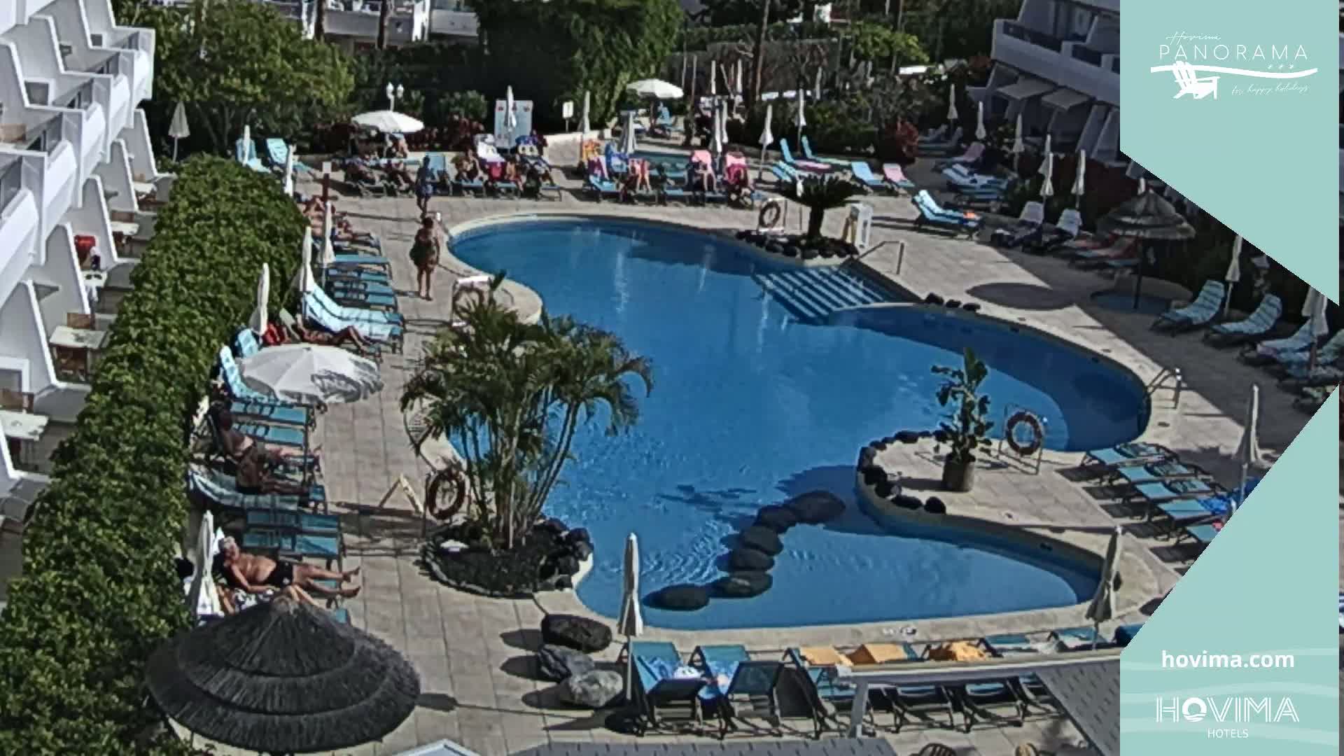 A sunny outdoor scene shows a kidney-shaped swimming pool surrounded by lounge chairs and umbrellas, with people relaxing and swimming.