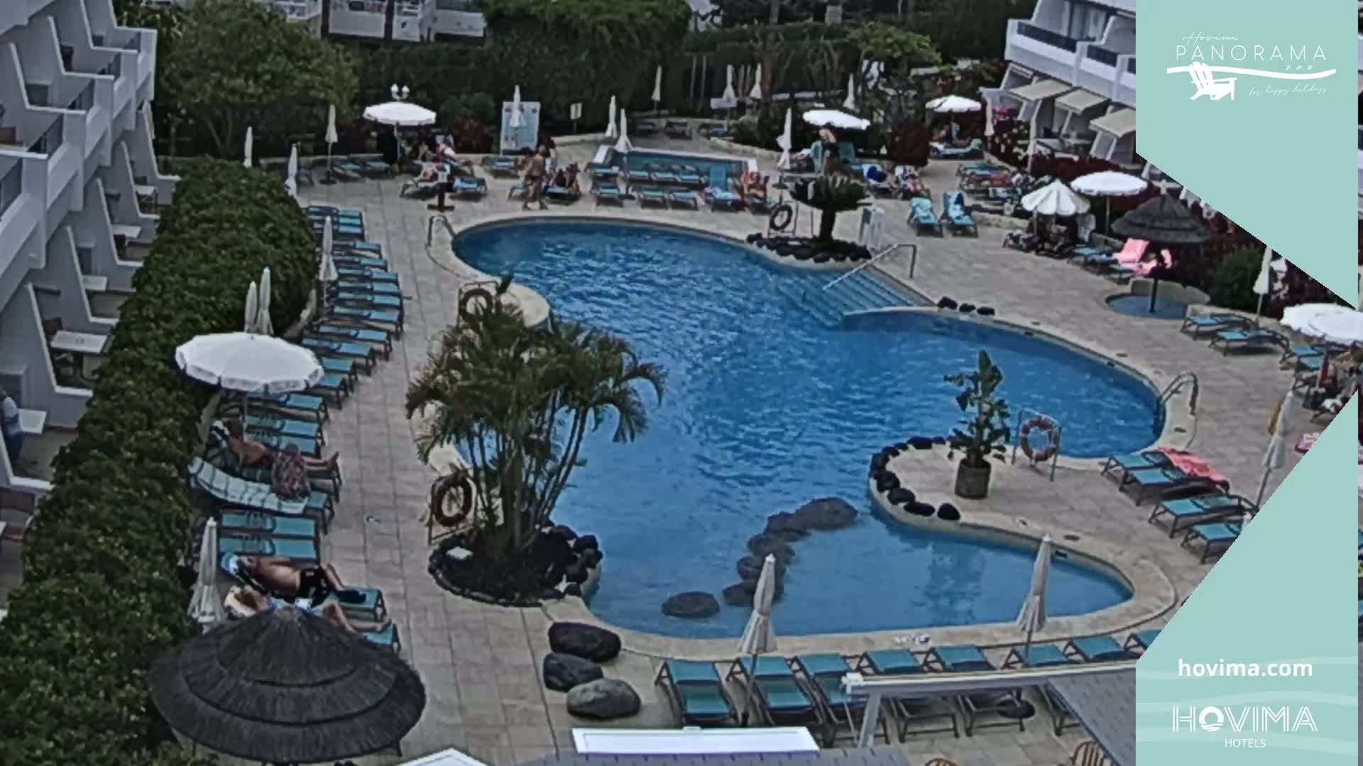An outdoor pool with surrounding lounge chairs and umbrellas sits next to a multi-story building on a partly sunny day.