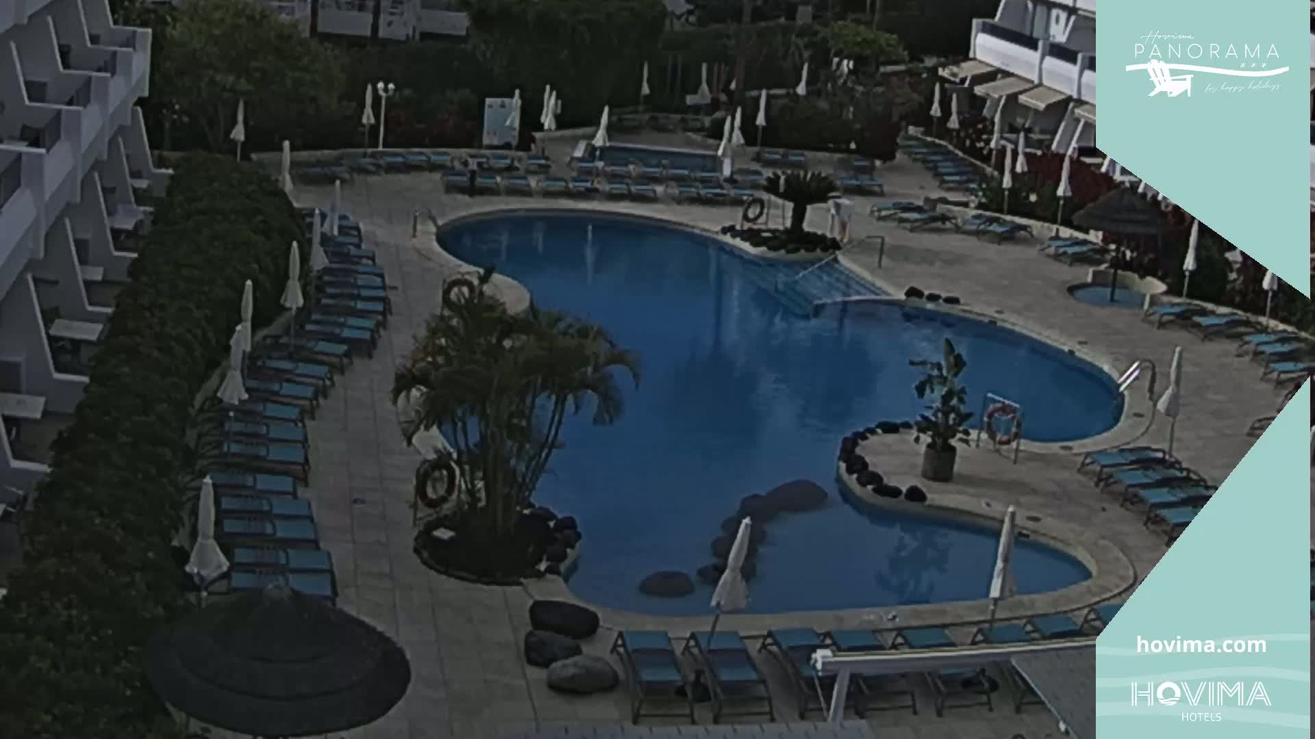 An outdoor swimming pool with surrounding lounge chairs and umbrellas is situated next to a building, under a cloudy sky.