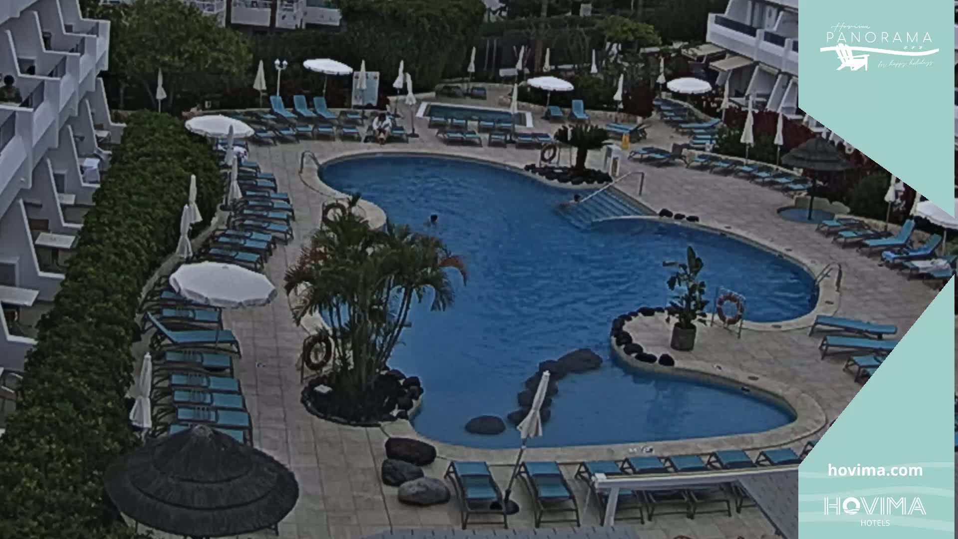 An outdoor pool with surrounding lounge chairs and umbrellas, situated next to a multi-story building on an overcast day.