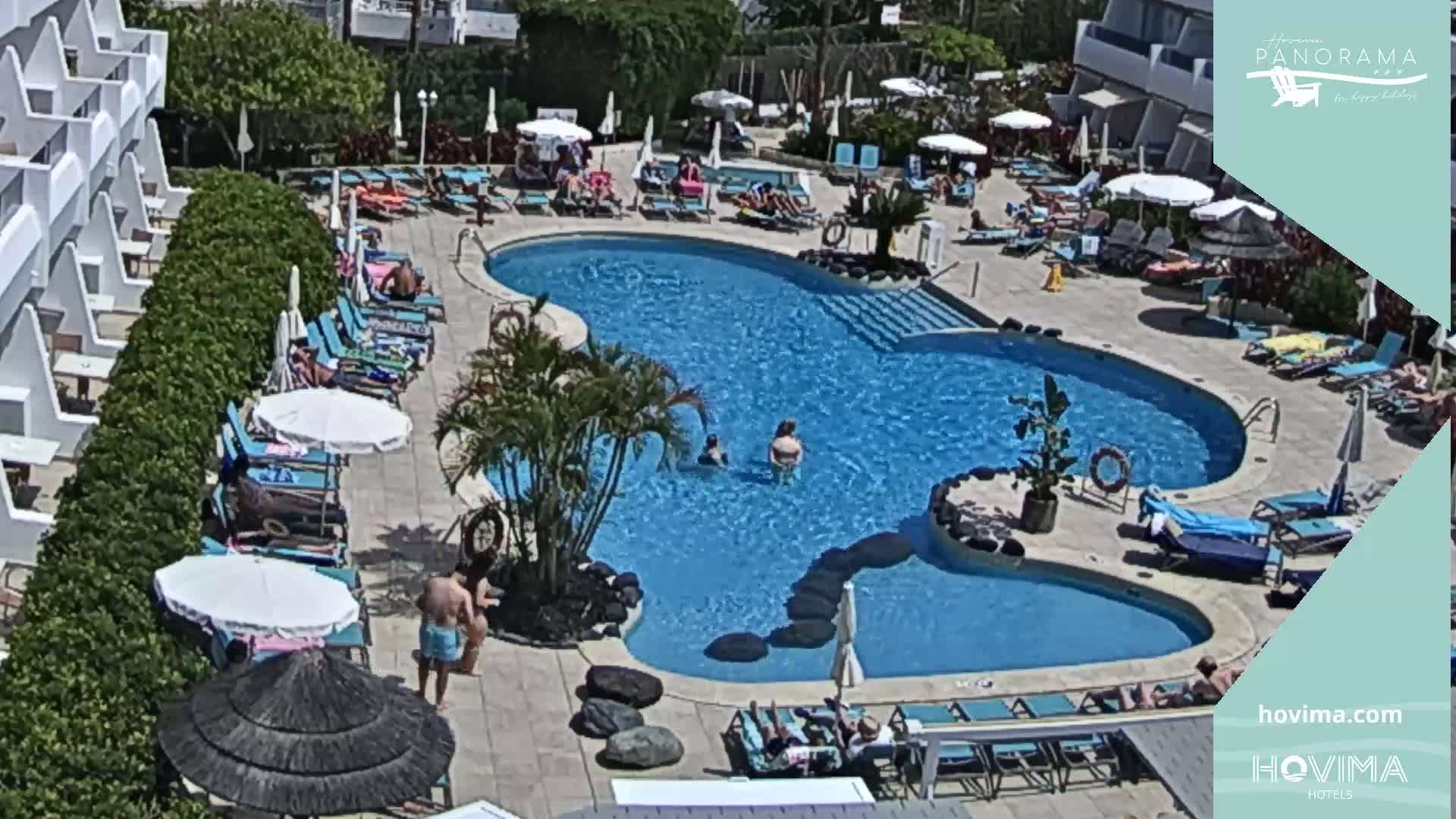 A sunny outdoor scene shows a kidney-shaped swimming pool surrounded by lounge chairs and umbrellas, with people relaxing and swimming.