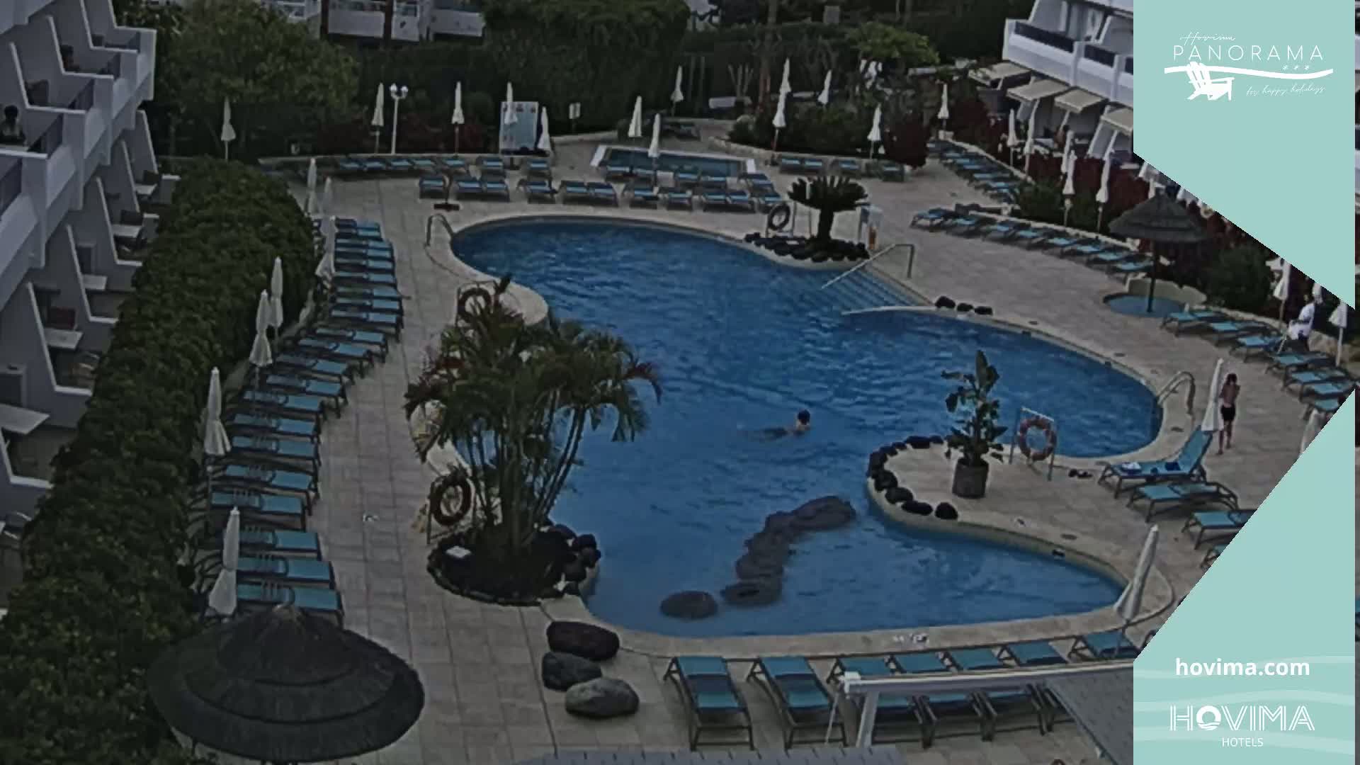 An outdoor pool surrounded by lounge chairs and umbrellas, next to a hotel building.