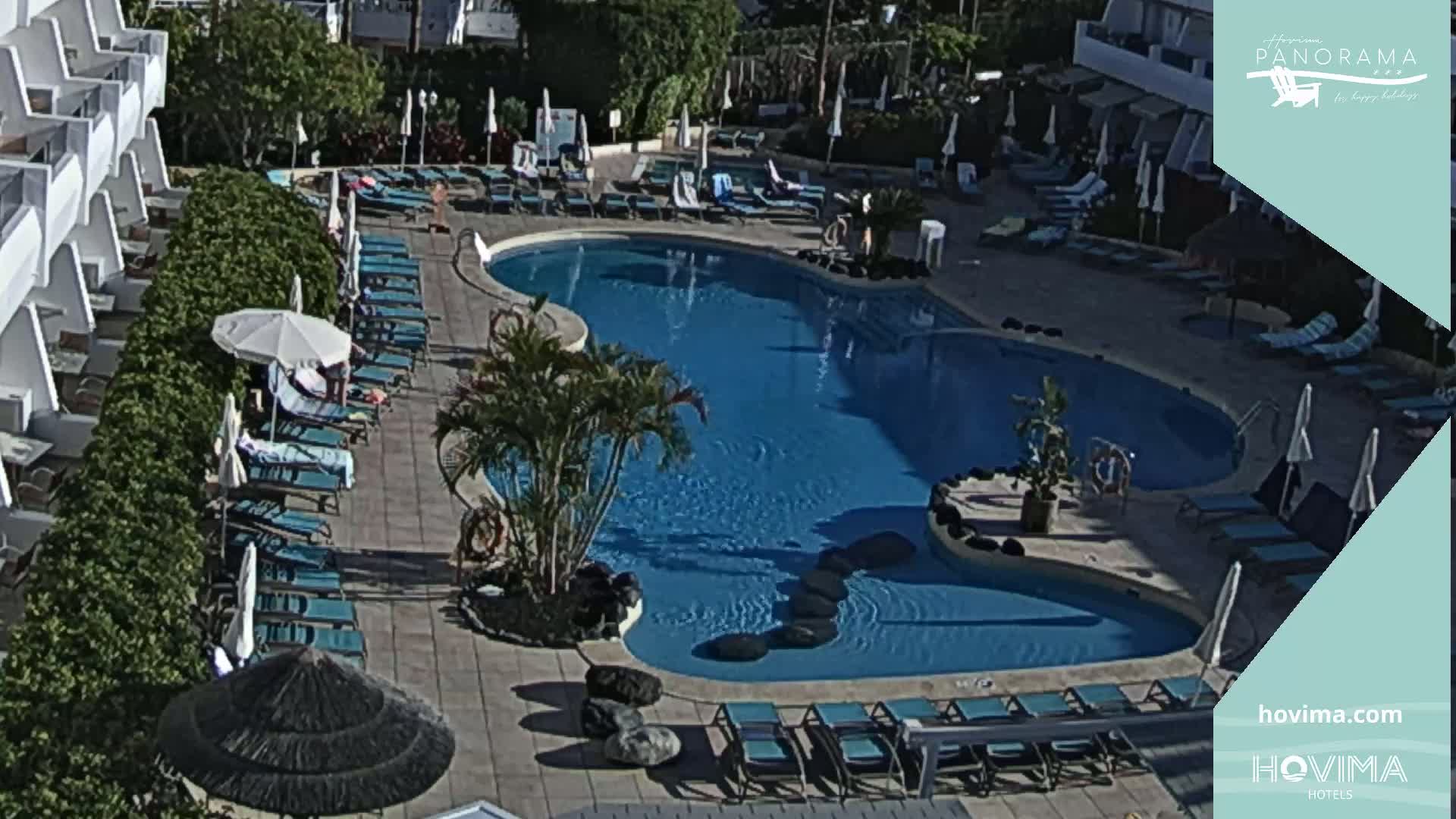 An outdoor swimming pool surrounded by lounge chairs and umbrellas, situated next to a multi-story building, on a sunny day.