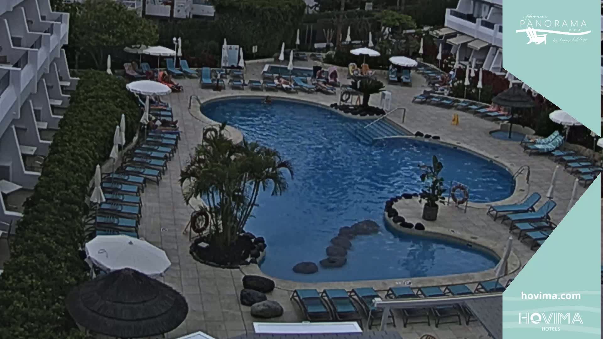 An outdoor swimming pool surrounded by lounge chairs and umbrellas, next to a hotel building on an overcast day.