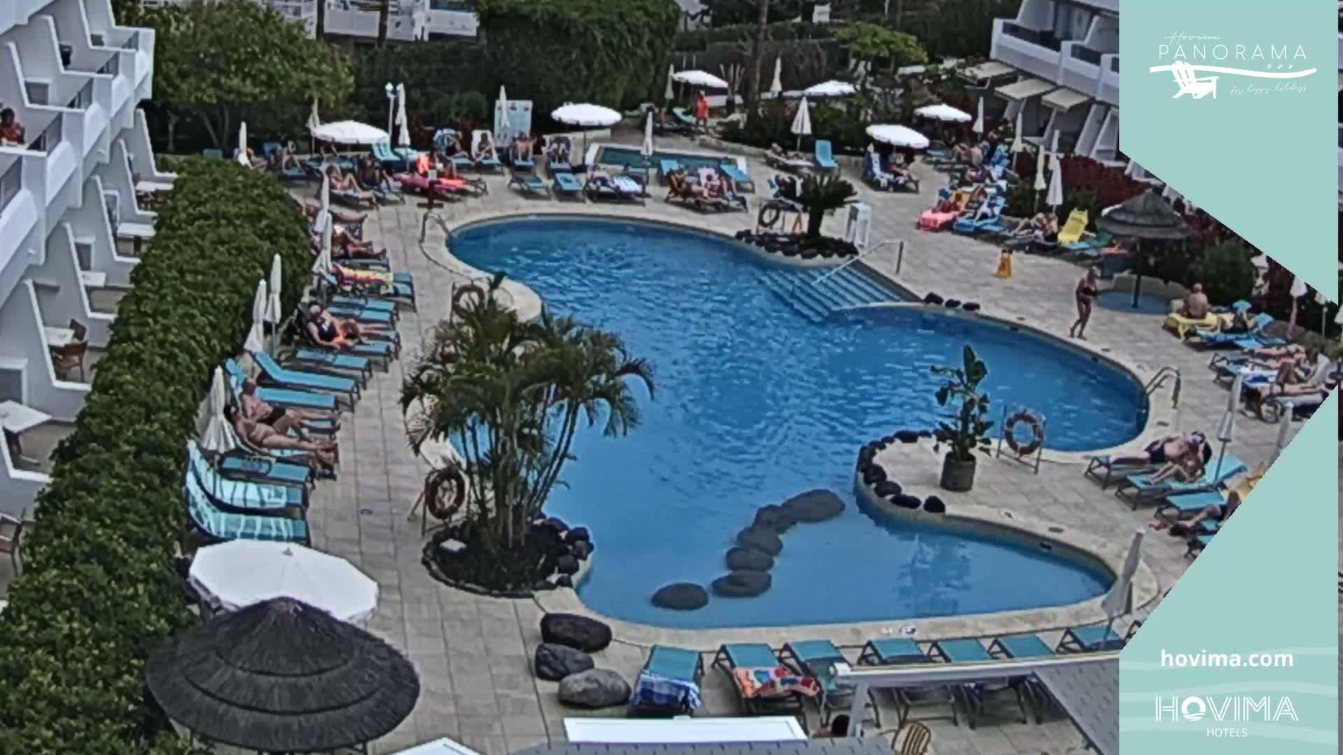 An outdoor pool with many people lounging on surrounding chairs and some in the water, under an overcast sky.
