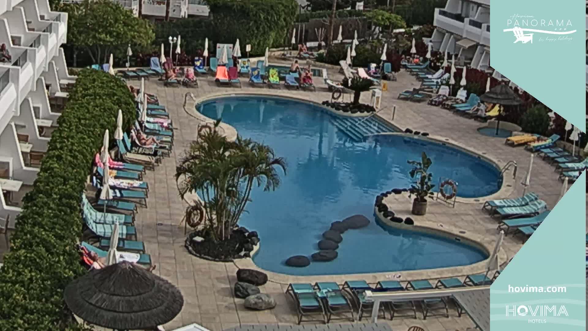An outdoor swimming pool surrounded by lounge chairs and umbrellas, with people relaxing around it on a sunny day.