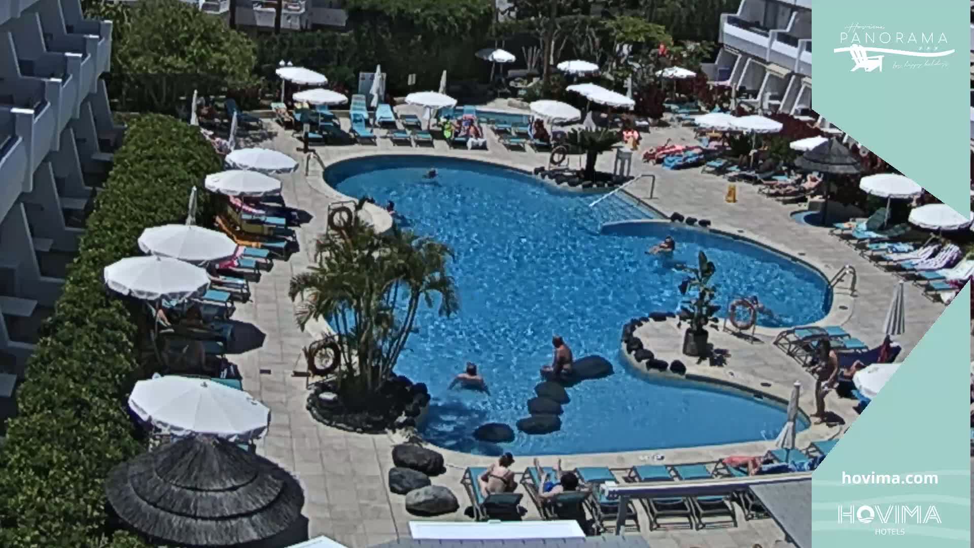 An outdoor swimming pool surrounded by lounge chairs, umbrellas, and buildings on a sunny day.