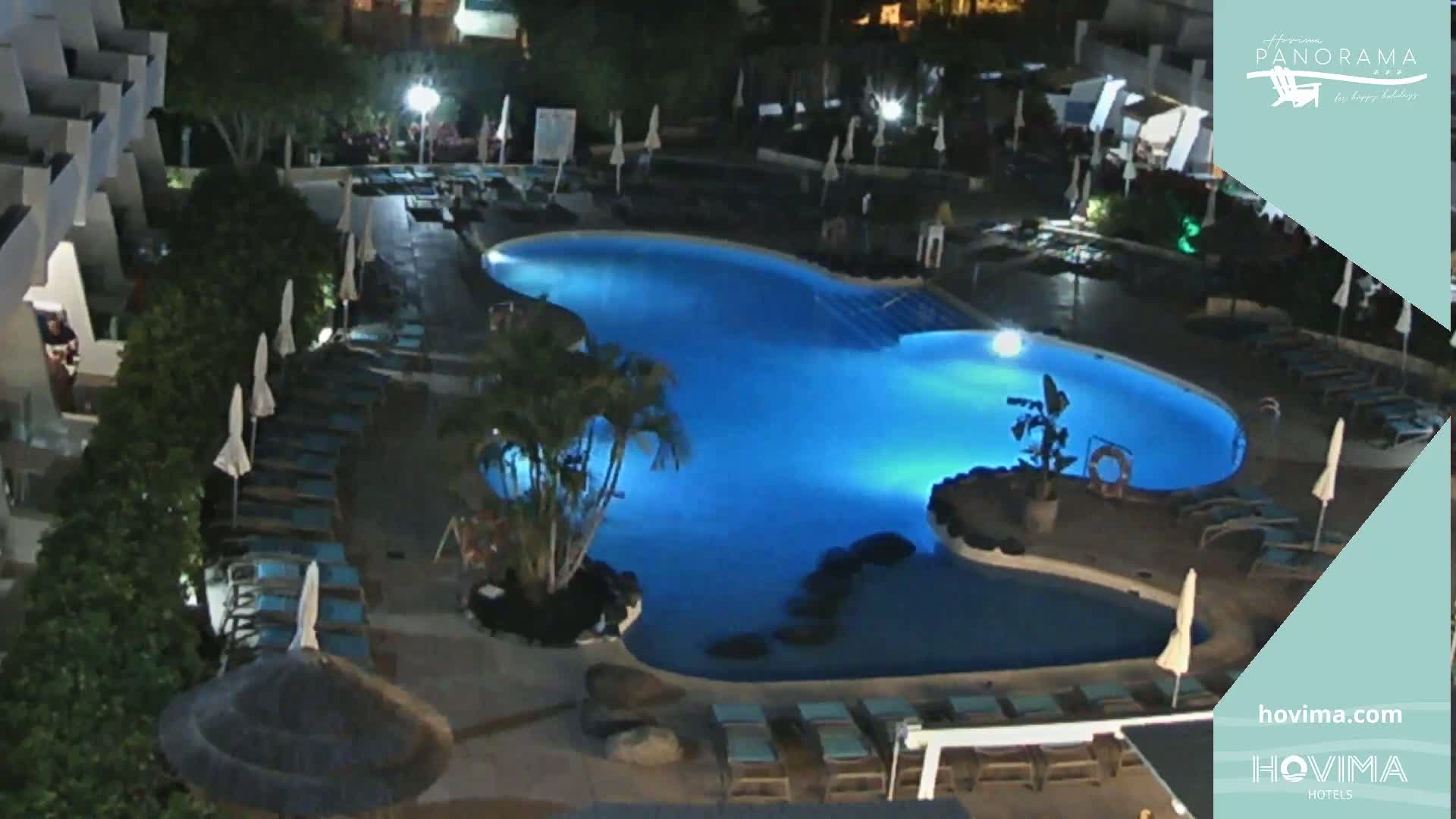 An illuminated swimming pool at night, surrounded by lounge chairs and umbrellas, sits within a resort complex under a dark sky.